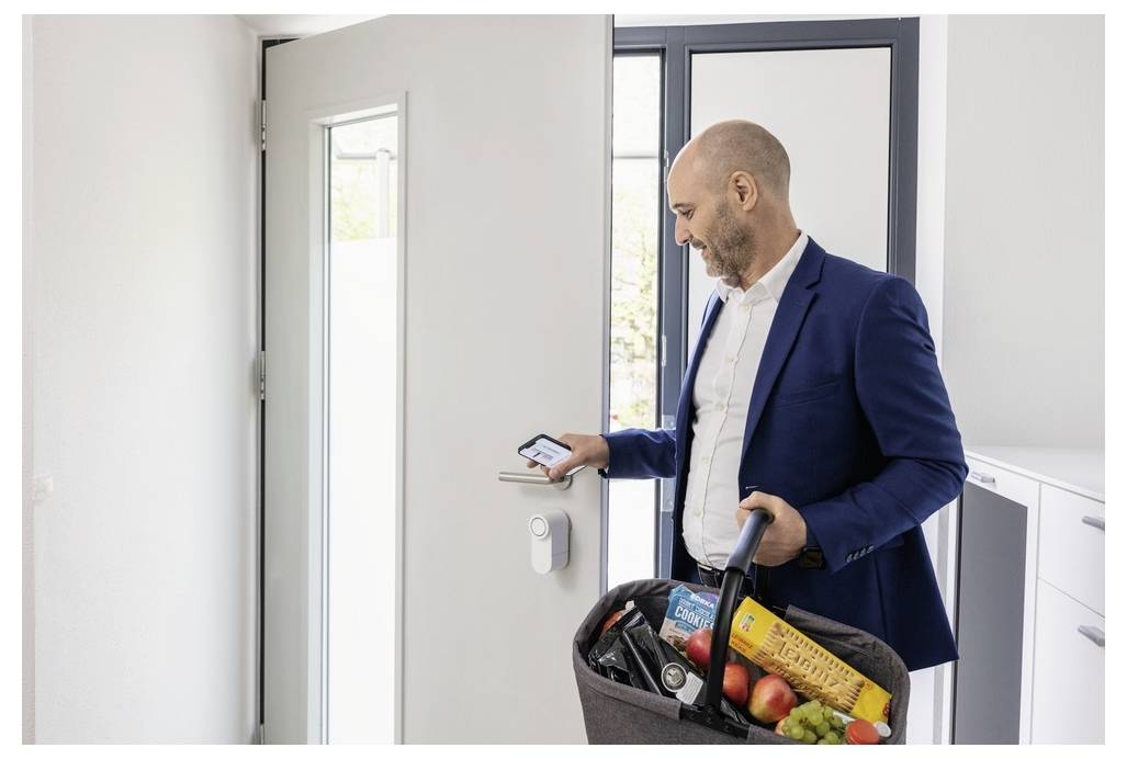 Une personne entre dans une maison avec un panier de courses, utilisant un smartphone pour déverrouiller une porte équipée d'une serrure intelligente.