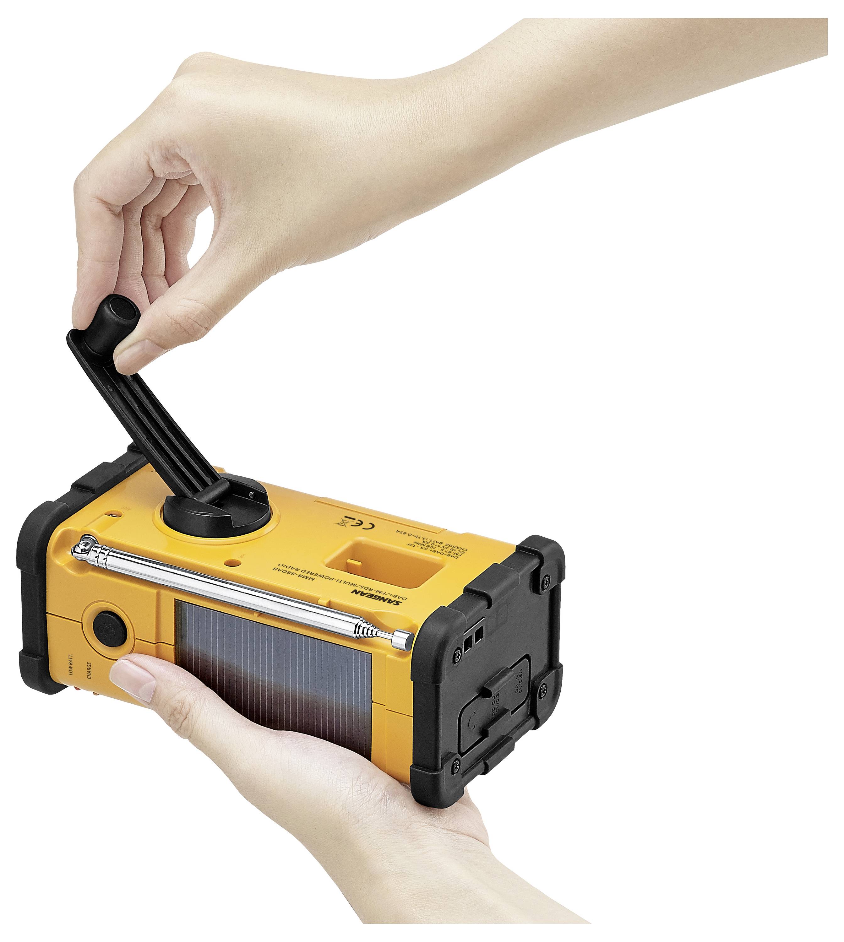 Une personne est en train de faire tourner manuellement un poste de radio d'urgence portable, jaune avec des coins en caoutchouc, équipé d'un panneau solaire et d'une lampe de poche sur le dessus.