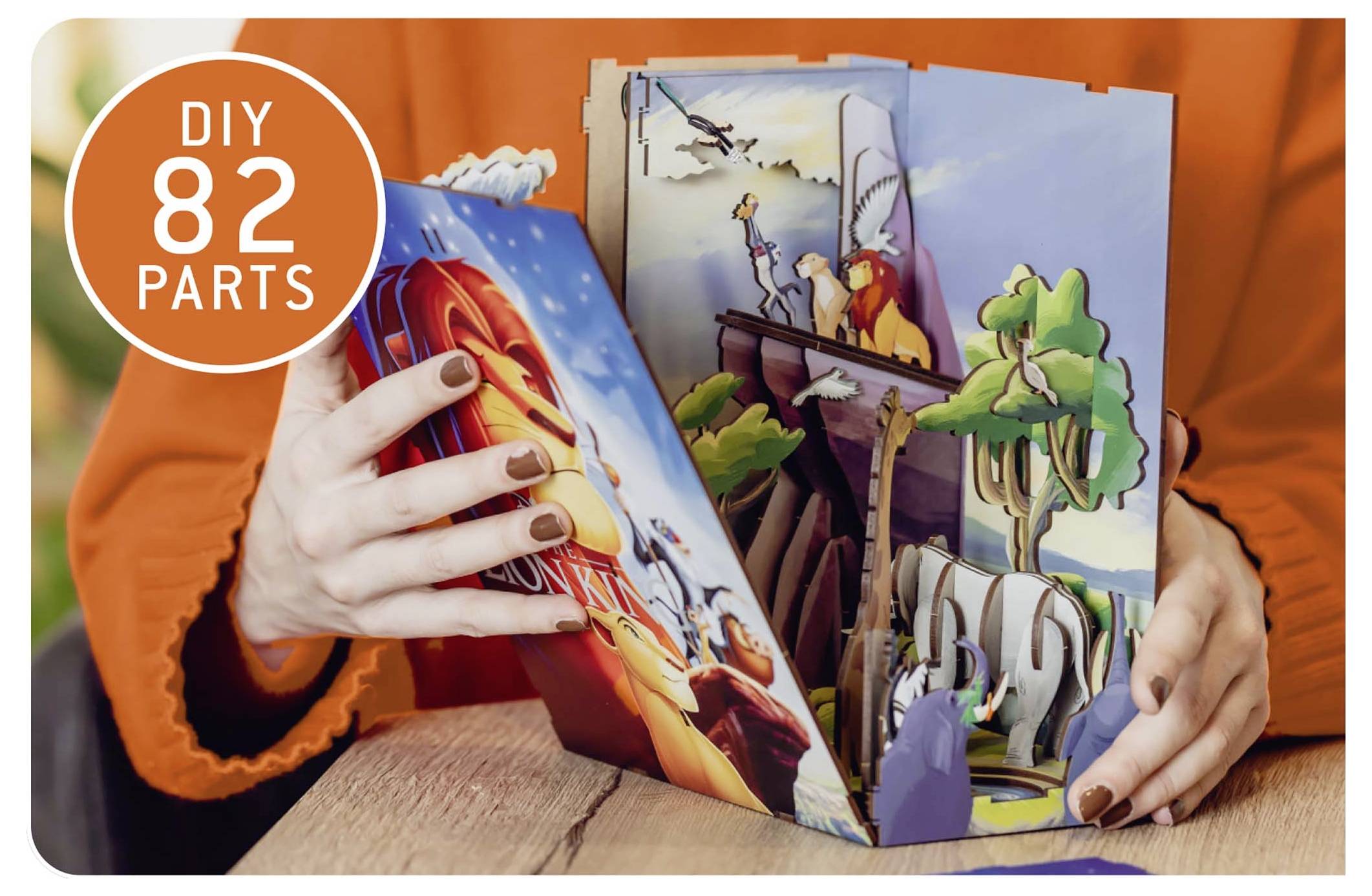 Une personne assemble un livre pop-up de bricolage coloré avec le texte « Bricolage 82 Pièces ». La scène représente des animaux et des paysages dans un diorama vibrant.