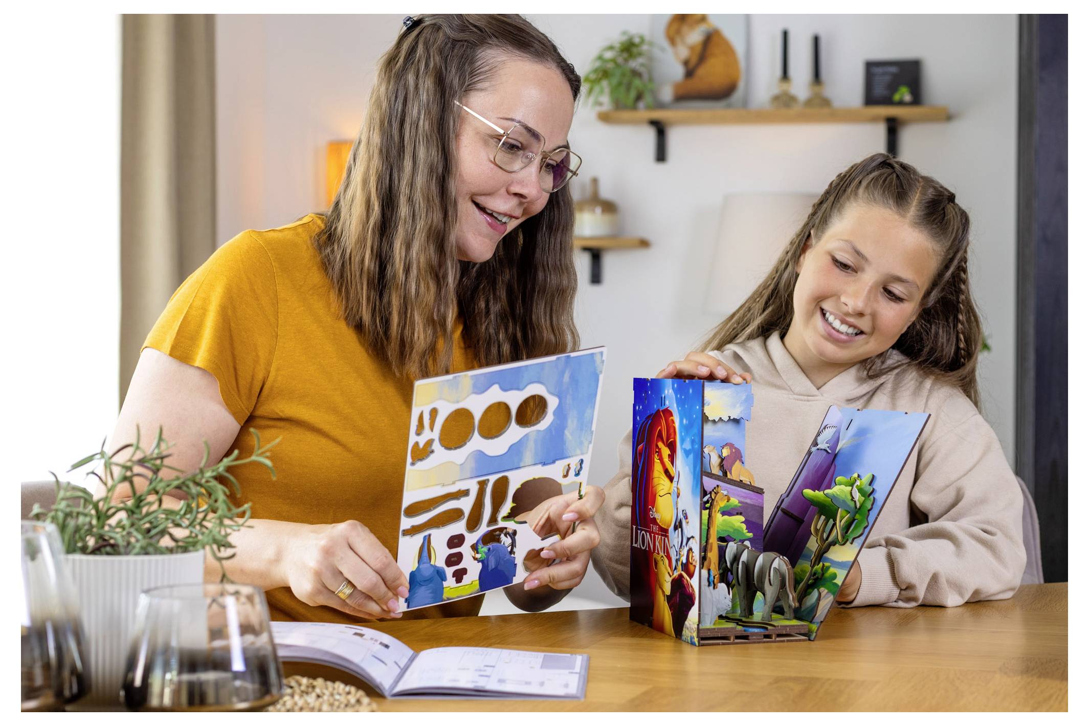 Une femme et un enfant sont assis à une table, souriant, tenant des livres illustrés ouverts. Une plante et divers objets sont posés sur la table.