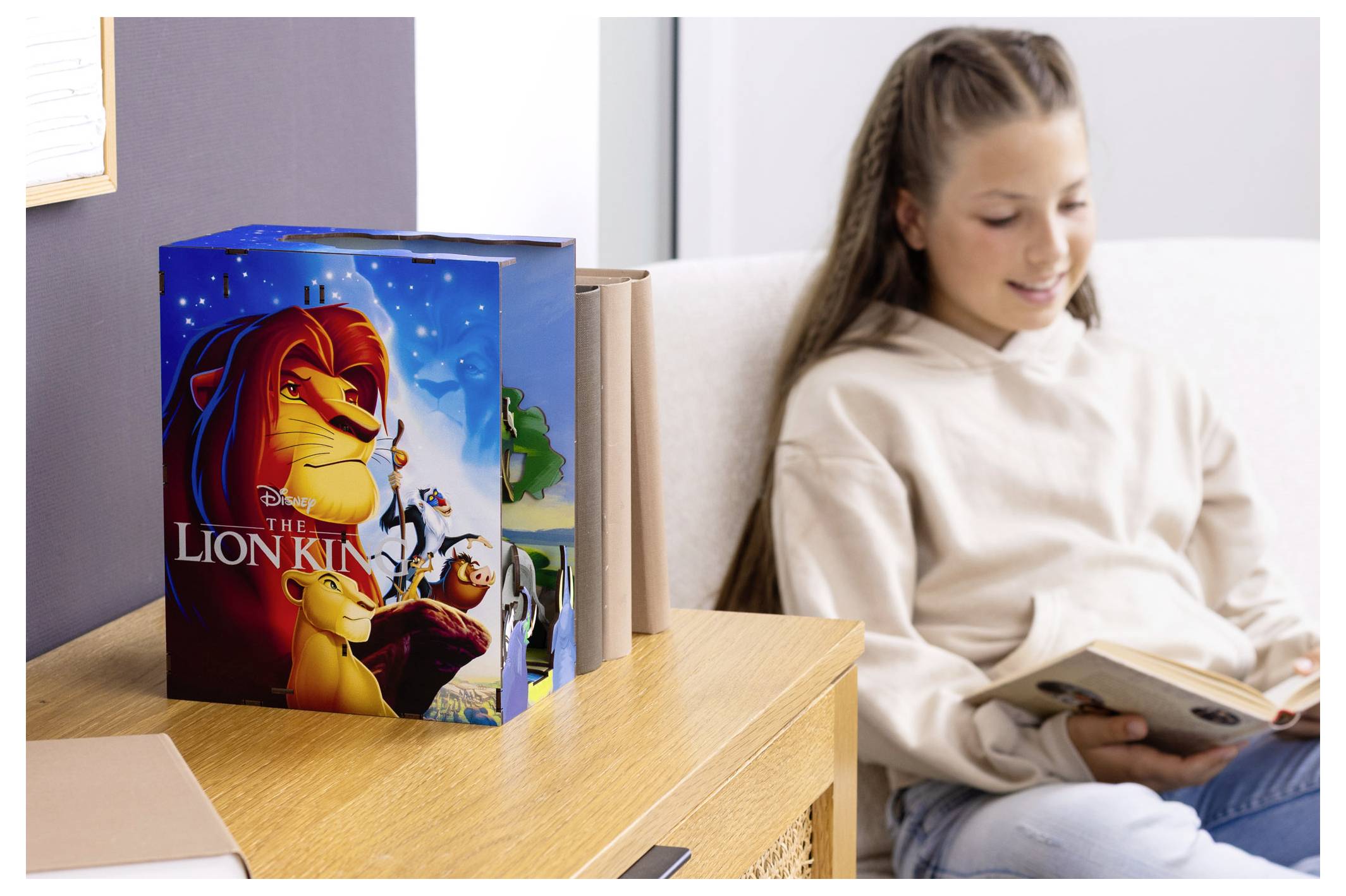Une jeune fille lit un livre à côté d'une boîte de jeu avec une illustration de 'Le Roi Lion' représentant ses personnages. La pièce dégage une atmosphère légère et chaleureuse.