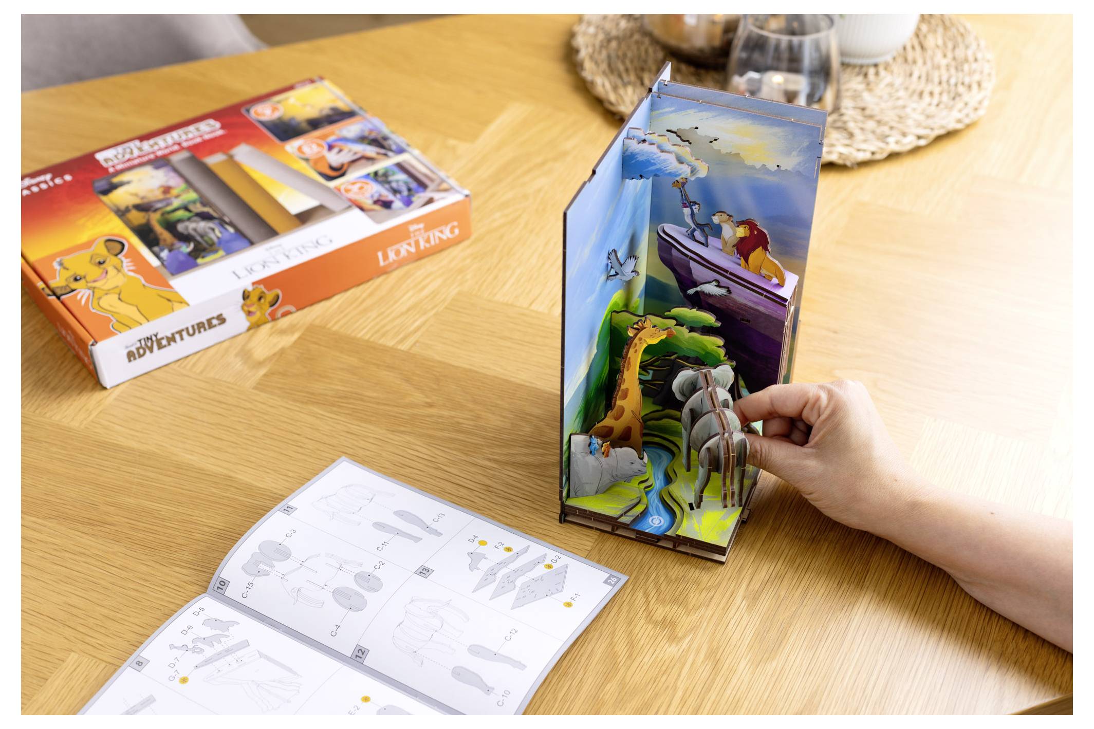 Une personne assemble un diorama en papier 3D d'une scène avec des animaux et un bateau, à côté d'un coffret de maquette sur le thème du Roi Lion et d'un livret d'instructions.
