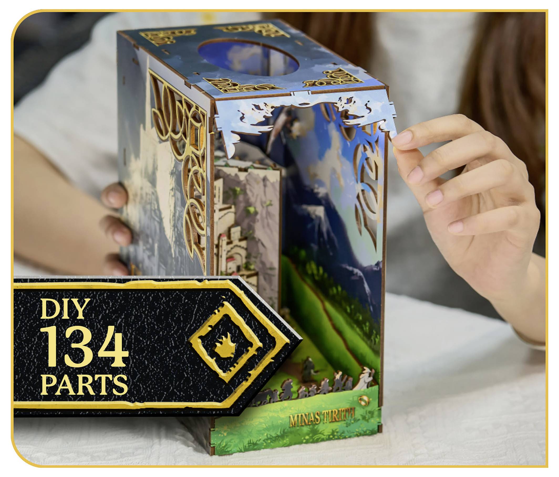 Kit de modèle de 134 pièces « À faire soi-même » présentant une scène détaillée de Minas Tirith avec une personne assemblant la structure, un arrière-plan pittoresque visible.