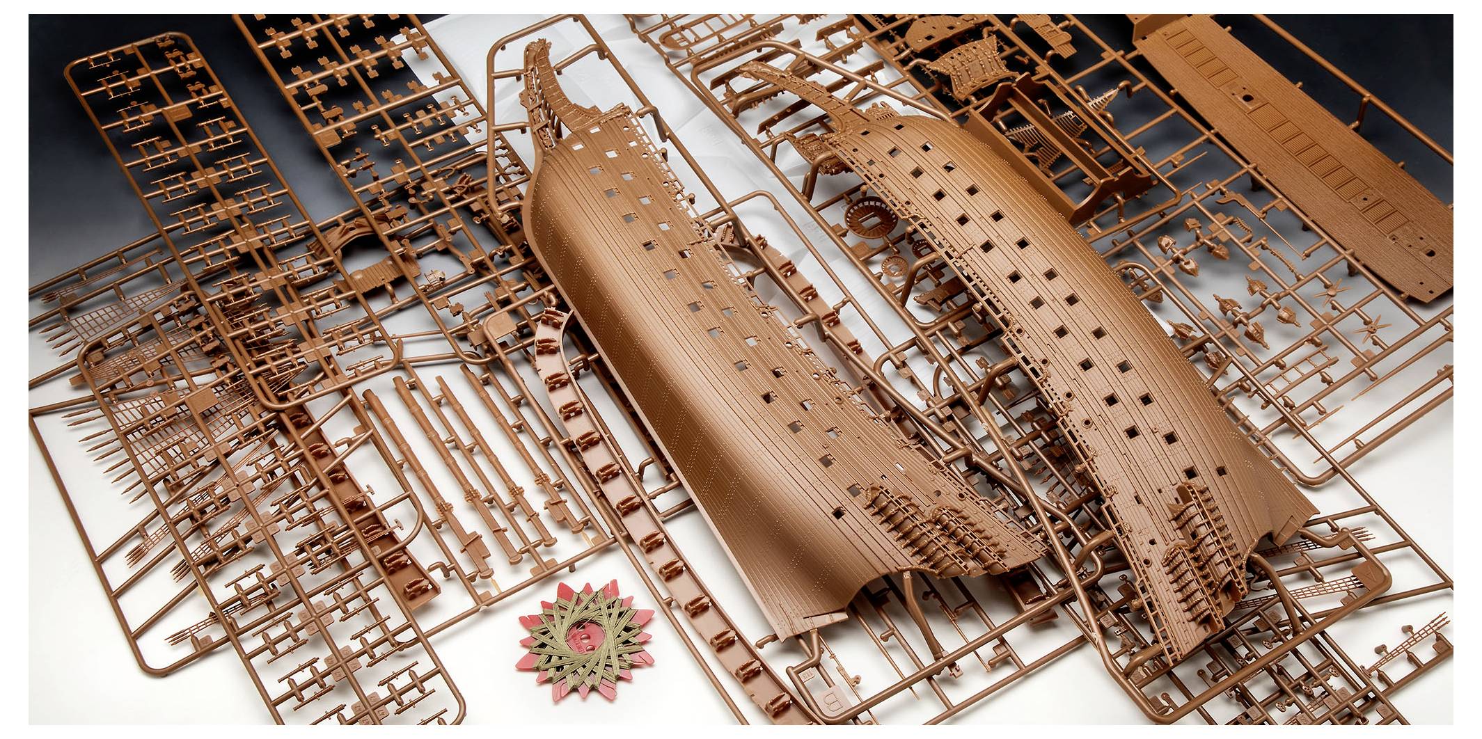 Feuille de pièces de maquette avec diverses pièces et une coque de navire partiellement assemblée en plastique marron, avec une pièce en forme de roue dentée au centre.
