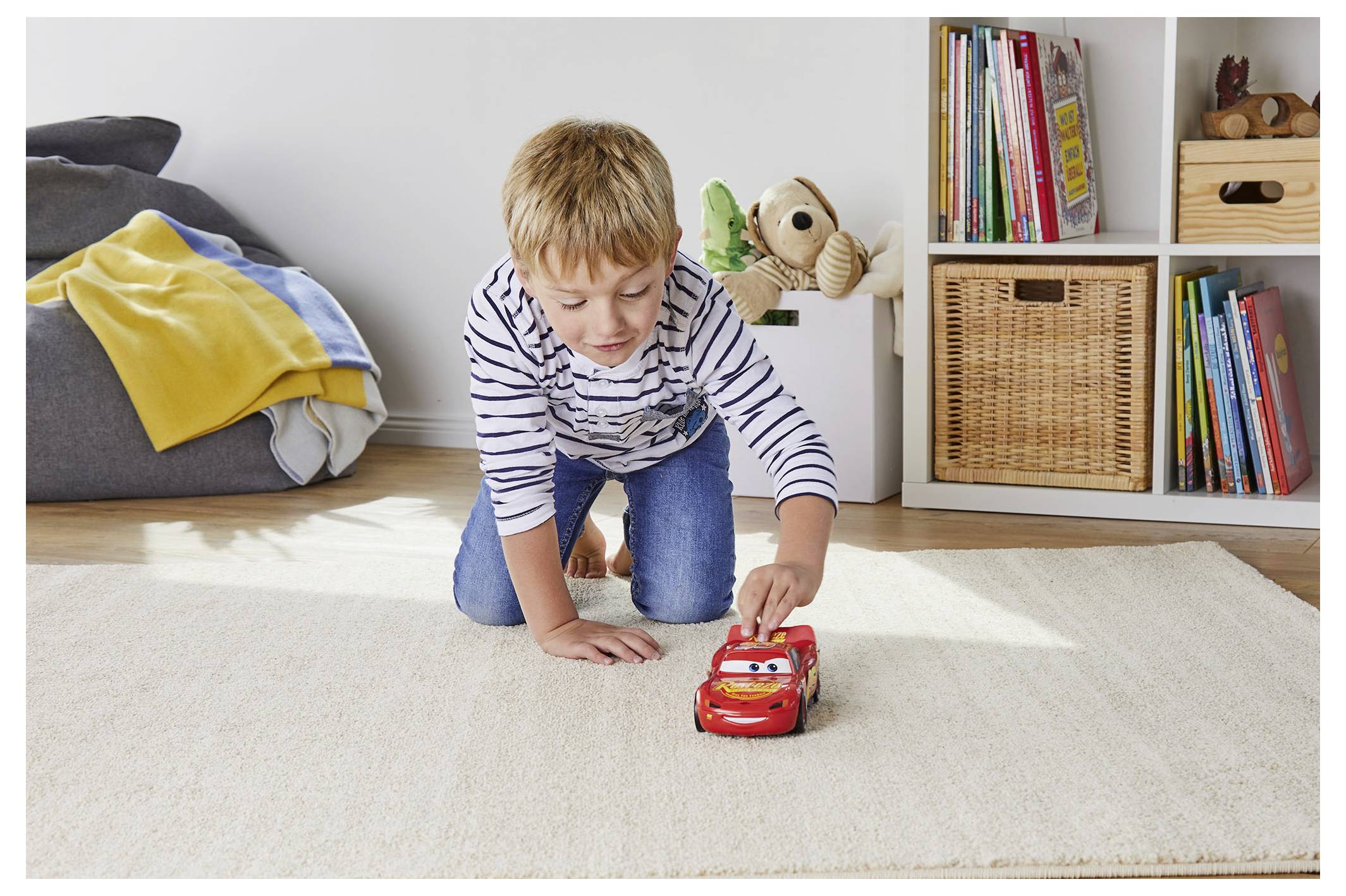 Un enfant joue sur un tapis avec une voiture miniature dans une pièce douillette, des étagères remplies de livres, un panier tressé et une pile de couvertures à proximité.