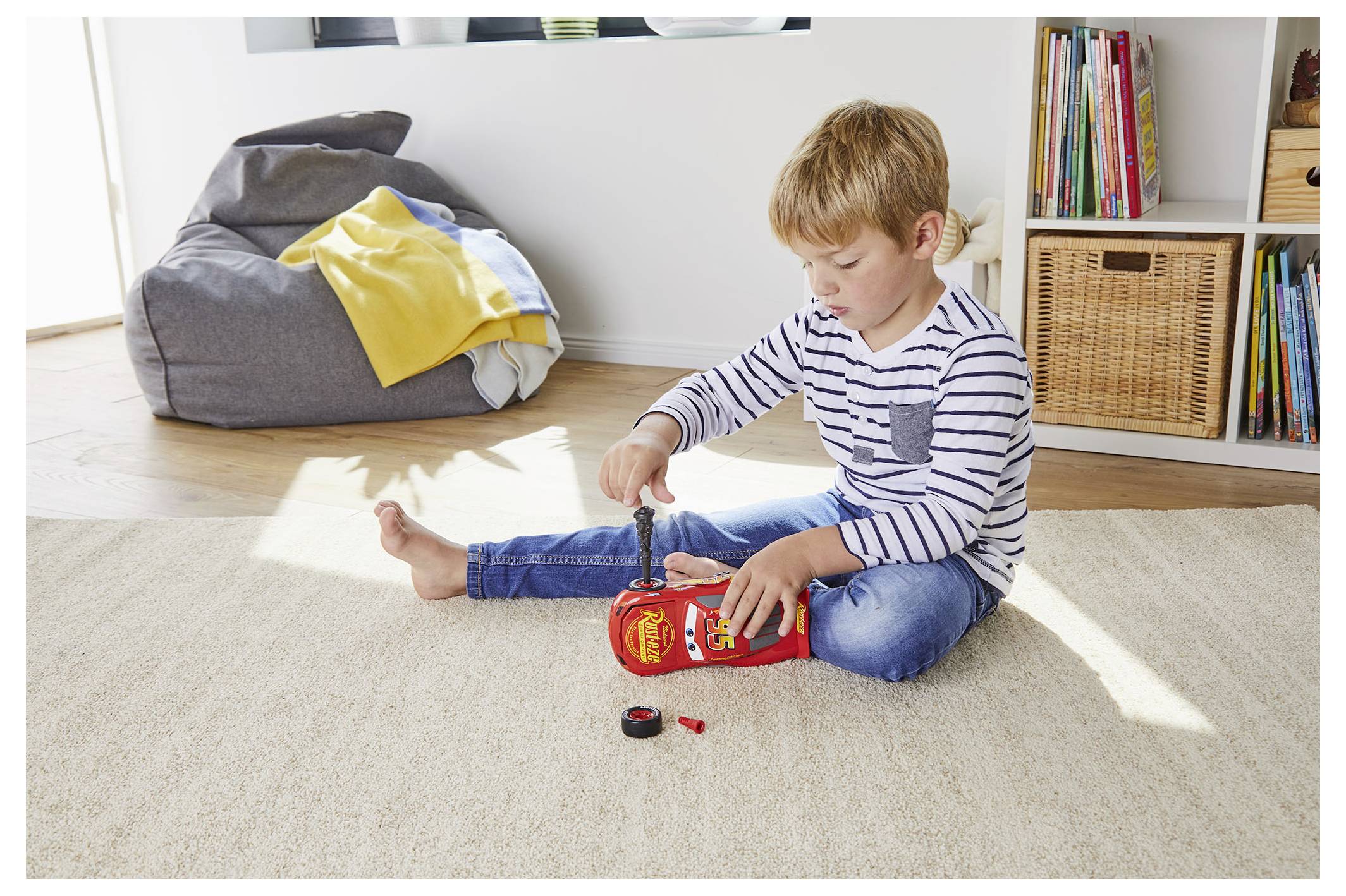 Un jeune enfant est assis sur un tapis, jouant avec une petite voiture dans une pièce bien éclairée, avec un pouf et une bibliothèque en arrière-plan.