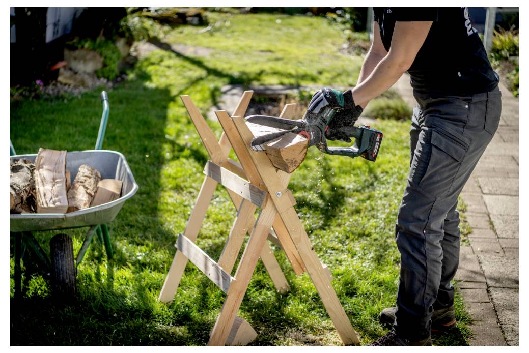 Une personne utilisant une tronçonneuse pour couper un rondin sur un chevalet de sciage dans un jardin. Une brouette remplie de bûches est proche, suggérant une activité de coupe de bois extérieure.