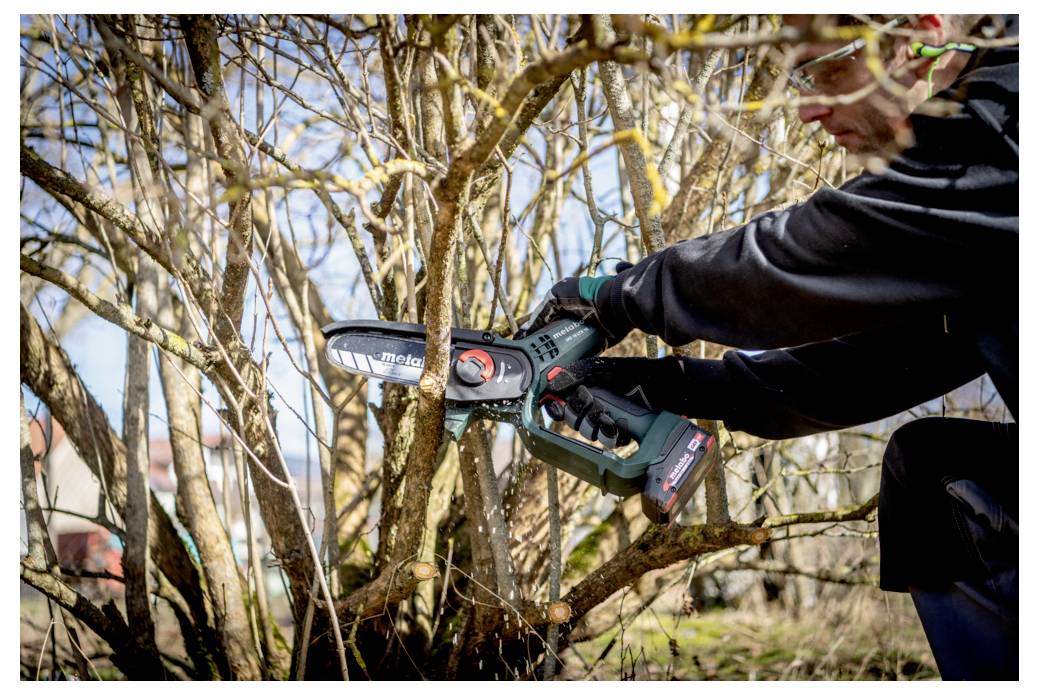 Une personne utilise une petite tronçonneuse électrique pour tailler des branches sur un arbre ou un arbuste dans un jardin, portant un équipement de protection.
