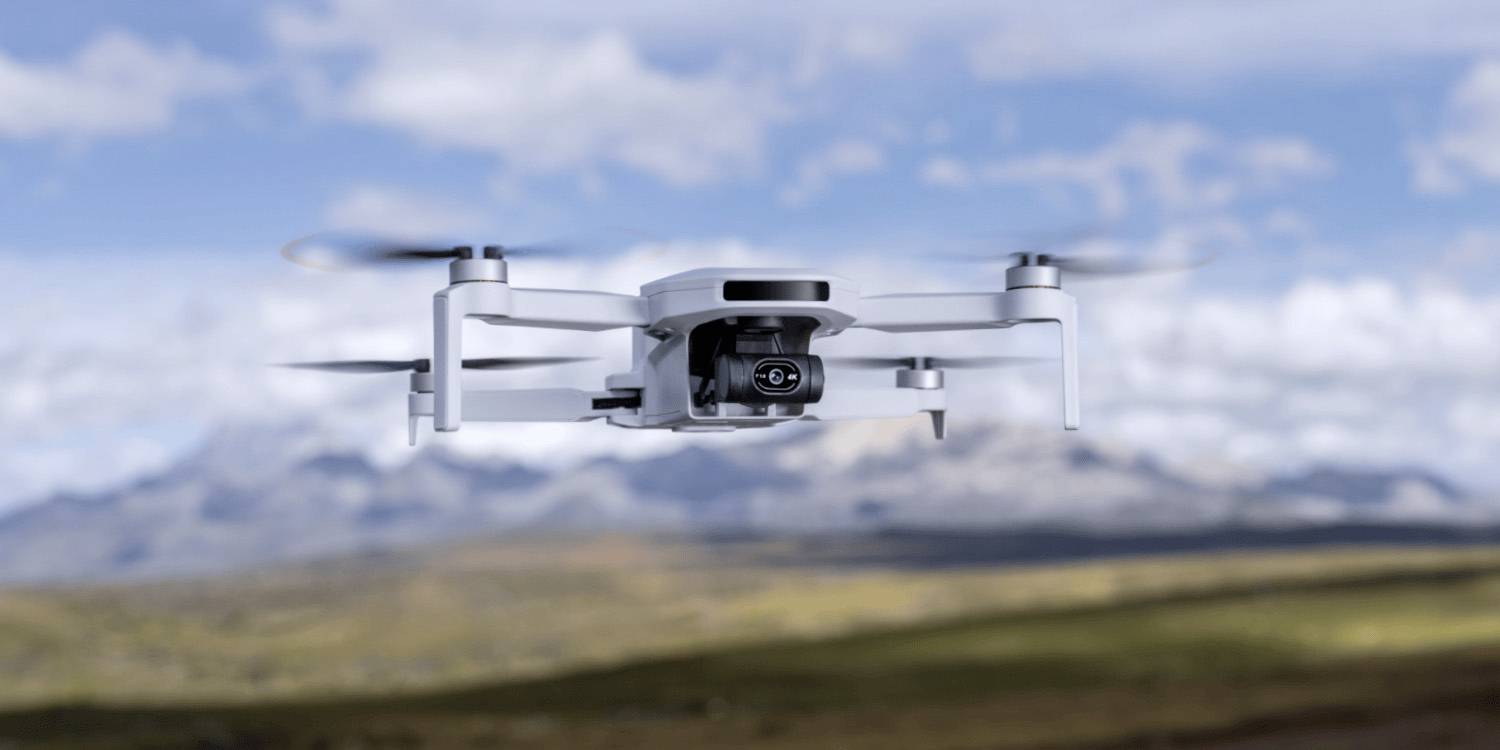 Un drone blanc équipé d'une caméra plane en premier plan, avec un paysage montagneux flou et un ciel bleu en arrière-plan.