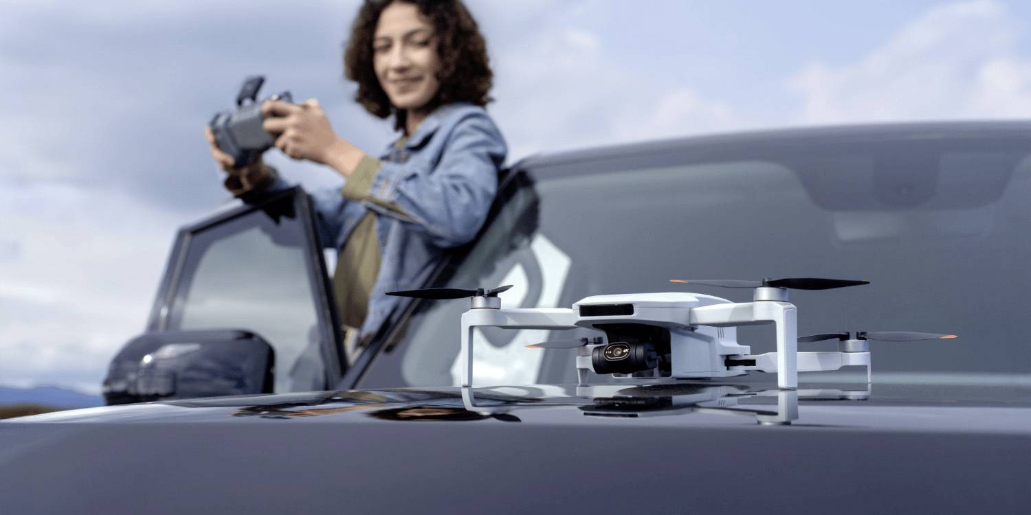 Une personne contrôle un drone avec une télécommande, debout à côté d'un SUV. Le drone est posé sur le capot du véhicule, prêt à voler.