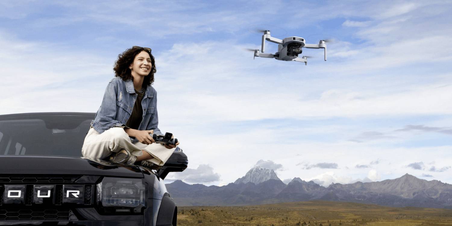 Une femme est assise sur un SUV noir 4x4, tenant une télécommande de drone. Un drone vole à proximité au-dessus d'un paysage pittoresque avec des montagnes en arrière-plan.