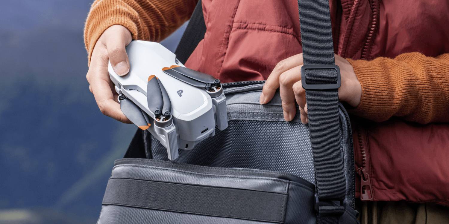 Une personne place un drone compact dans un sac à bandoulière, suggérant sa portabilité et sa facilité d'utilisation. La personne porte une veste rouge et des manches orange.