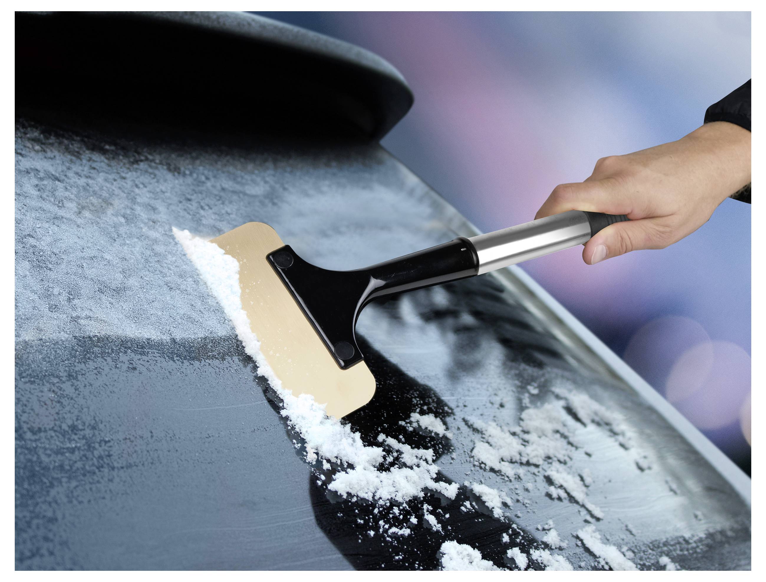 Plan rapproché d'une main utilisant un grattoir pour enlever la glace du pare-brise d'une voiture, dévoilant la vitre transparente en dessous.