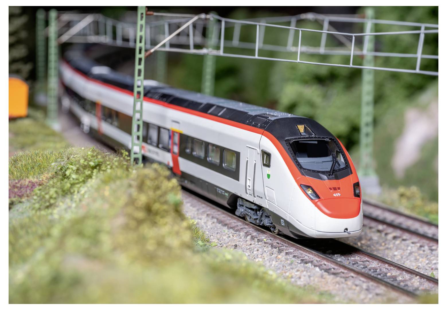 Un train miniature aux couleurs rouge et blanc circule sur des rails sous des portiques métalliques verts, entouré d'un paysage miniature détaillé.