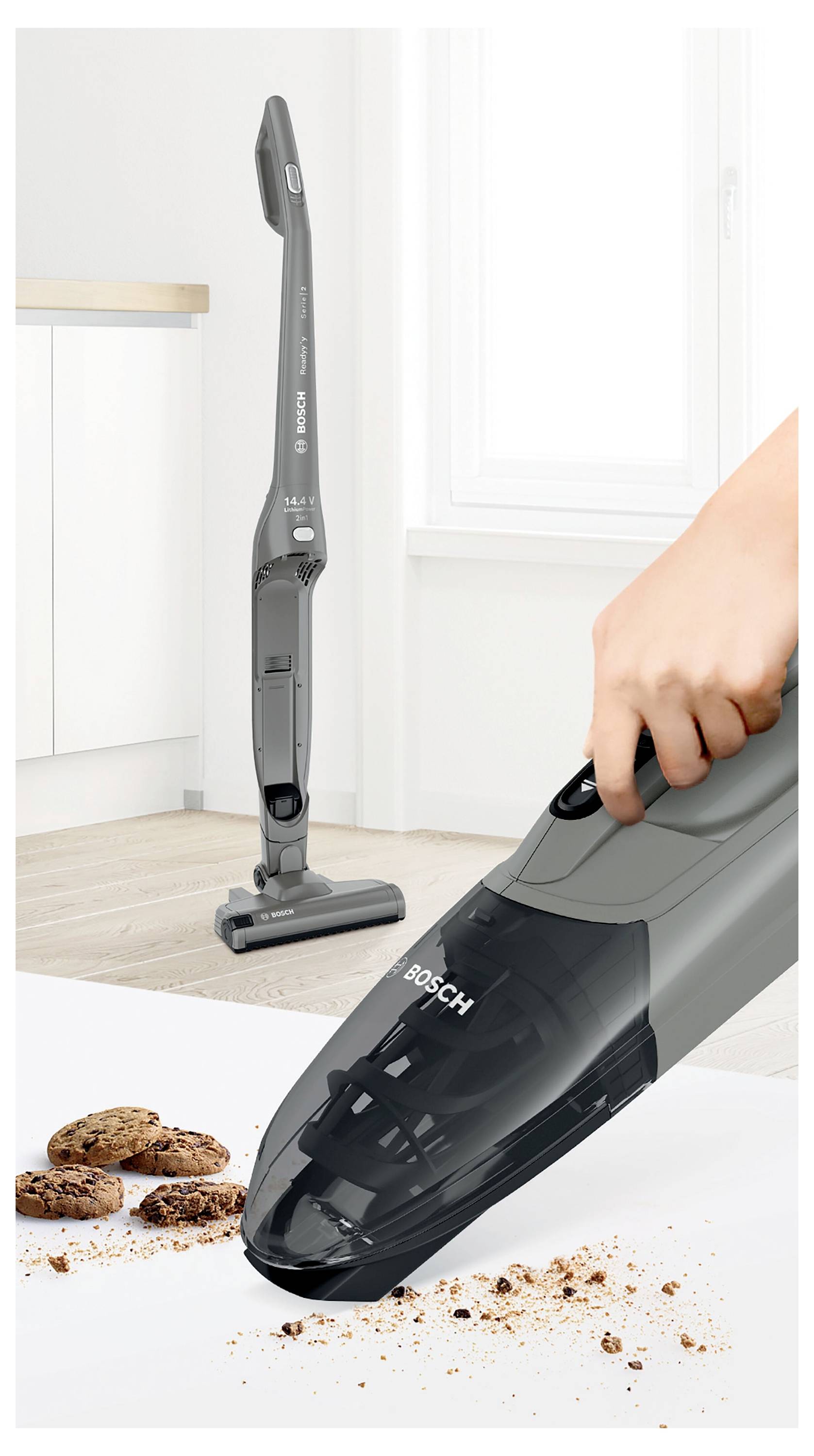Une main utilise un aspirateur Bosch pour ramasser des miettes de cookies sur une surface blanche. Un autre aspirateur est dressé verticalement en arrière-plan.
