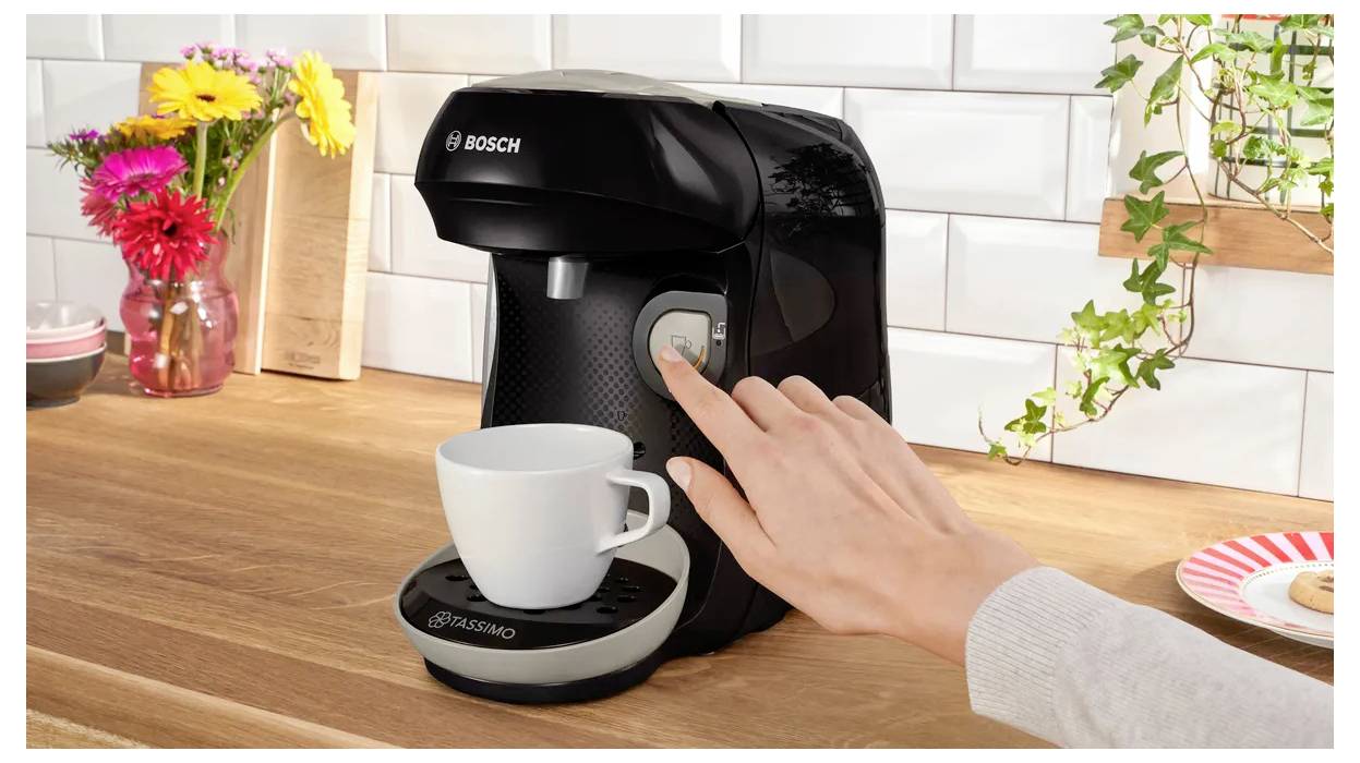 Une personne appuie sur un bouton d'une machine à café noire ; une tasse blanche attend en dessous du distributeur. Des fleurs colorées et des tasses sont en arrière-plan.