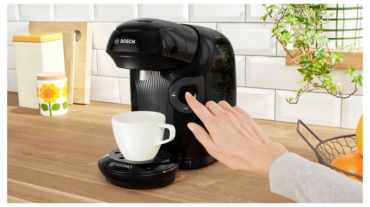 Une main appuie sur un bouton d'une machine à café noire Bosch. Une tasse blanche est posée sur le support. Plan de travail de cuisine avec des éléments de décoration en arrière-plan.