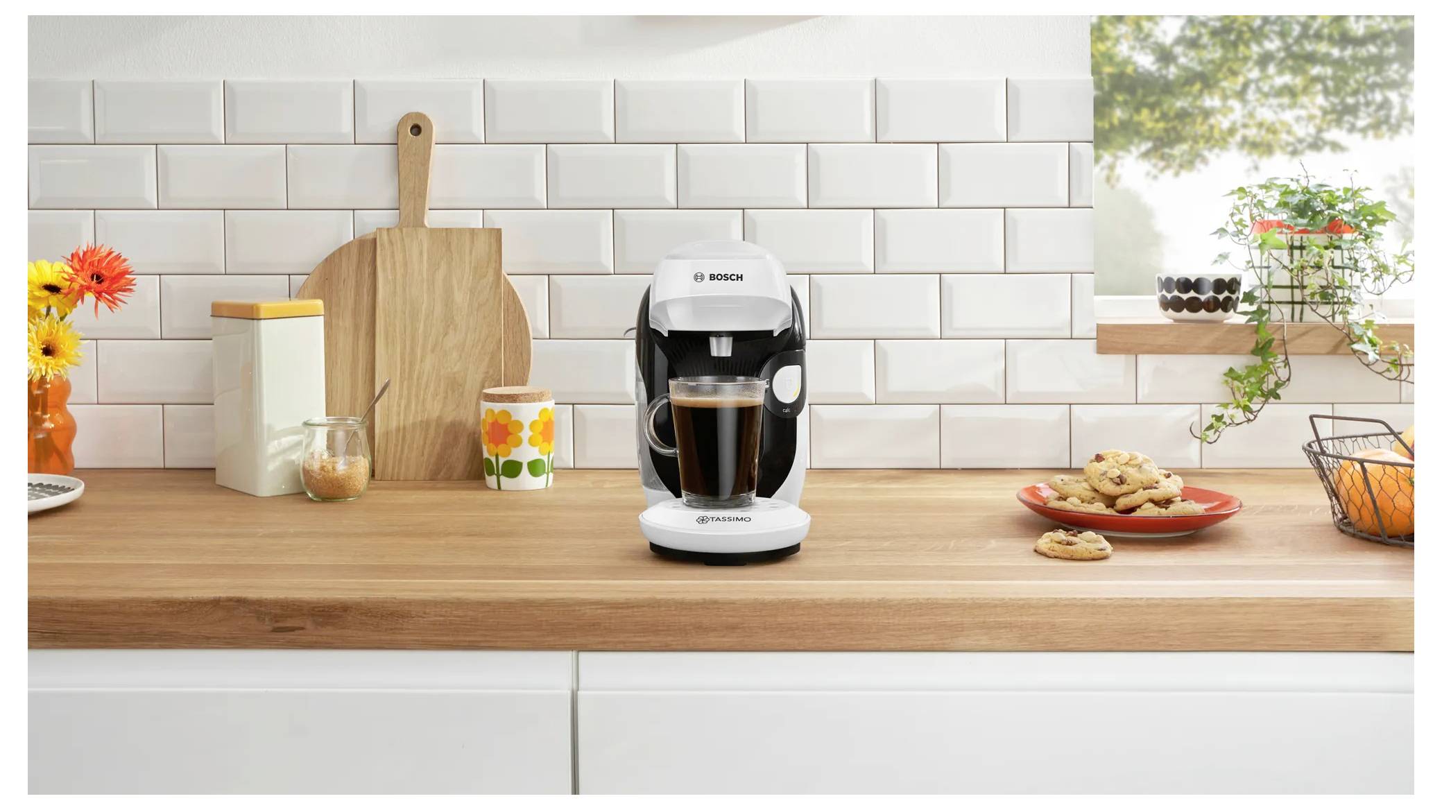 Un cafetière moderne préparant une tasse sur un comptoir de cuisine en bois ; entourée de cookies, de fleurs et de bocaux contre un mur de carreaux blancs.