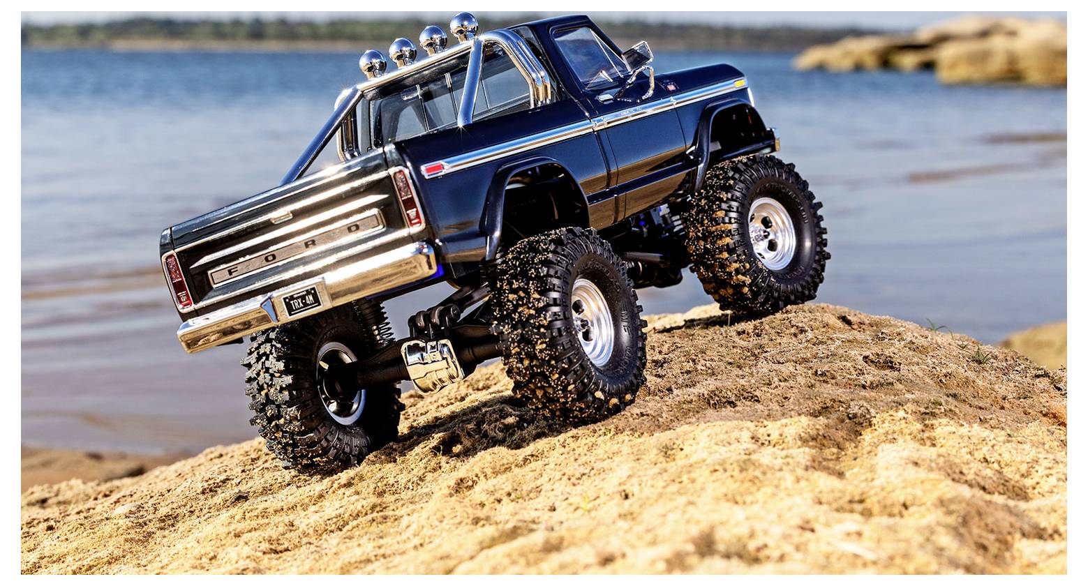 Un camion tout-terrain télécommandé bleu, doté de grandes roues, gravit une colline sablonneuse au bord d'un lac, mettant en valeur son design robuste et ses capacités.
