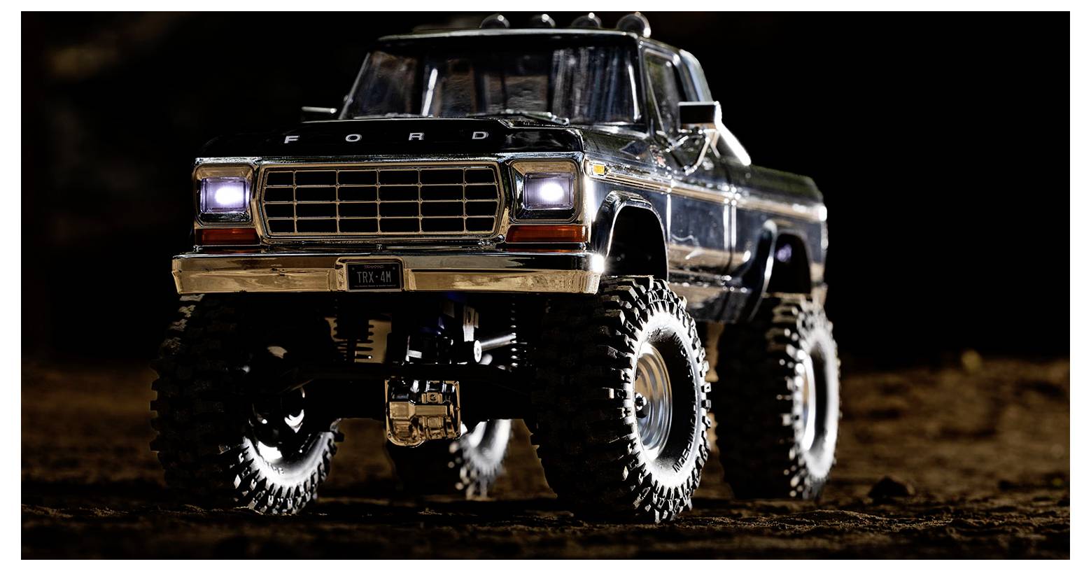 Un camion jouet avec de grandes roues tout-terrain et ses phares allumés, garé sur une surface terreuse pendant la nuit.