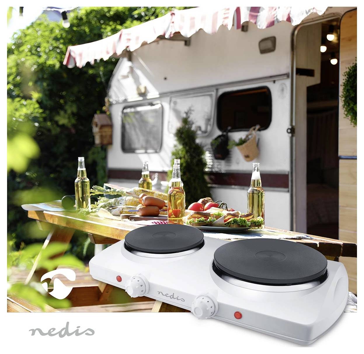 Une double plaque chauffante sur une table en bois, avec des aliments et des boissons, installée à l'extérieur d'une caravane, avec de la verdure et un auvent en arrière-plan.