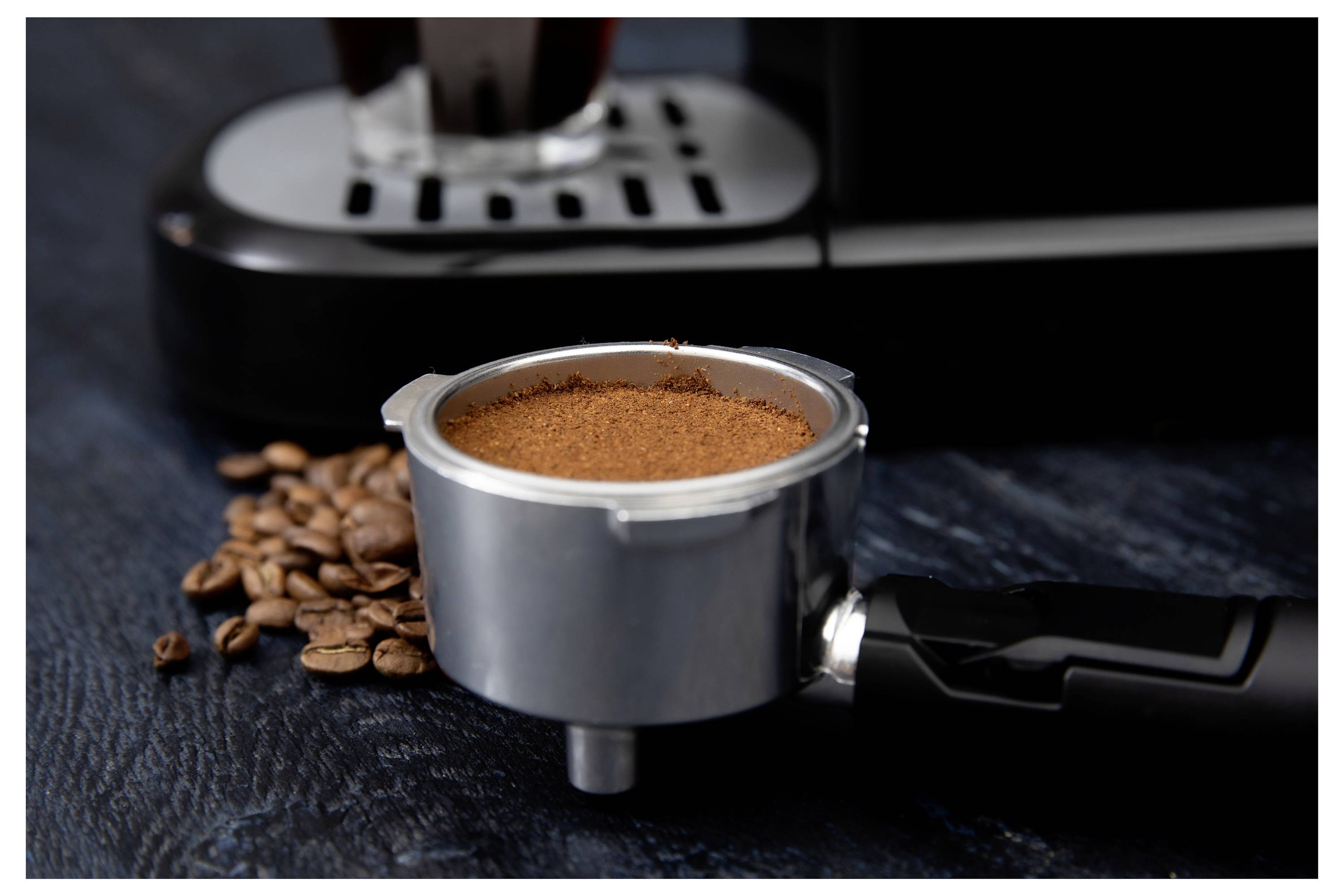 Portafilter rempli de café moulu à côté de grains de café, placé devant une machine à espresso sur un fond sombre.