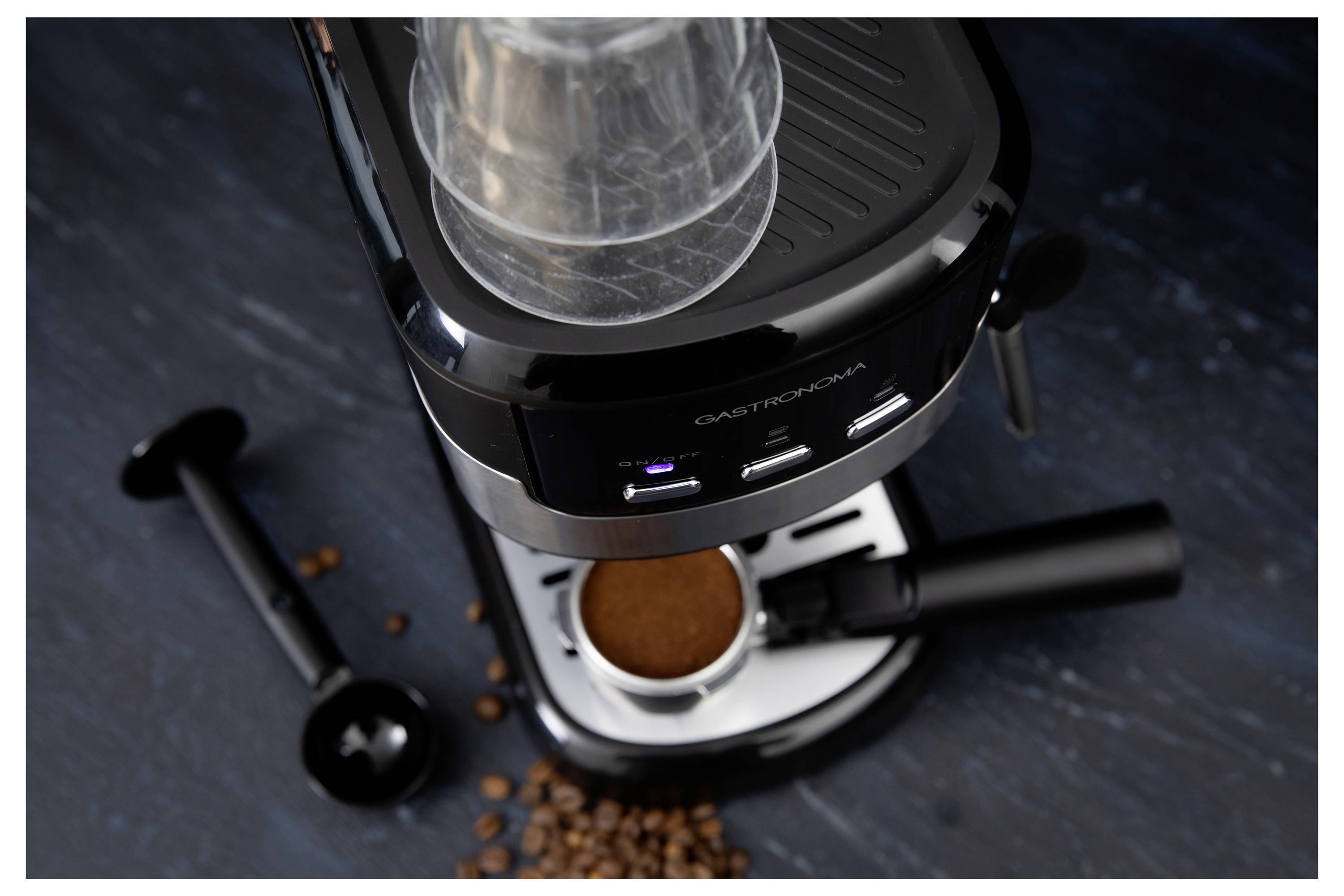 Une machine à espresso noire avec du café moulu dans le porte-filtre, entourée de grains de café et d'un tamper, sur une surface sombre.
