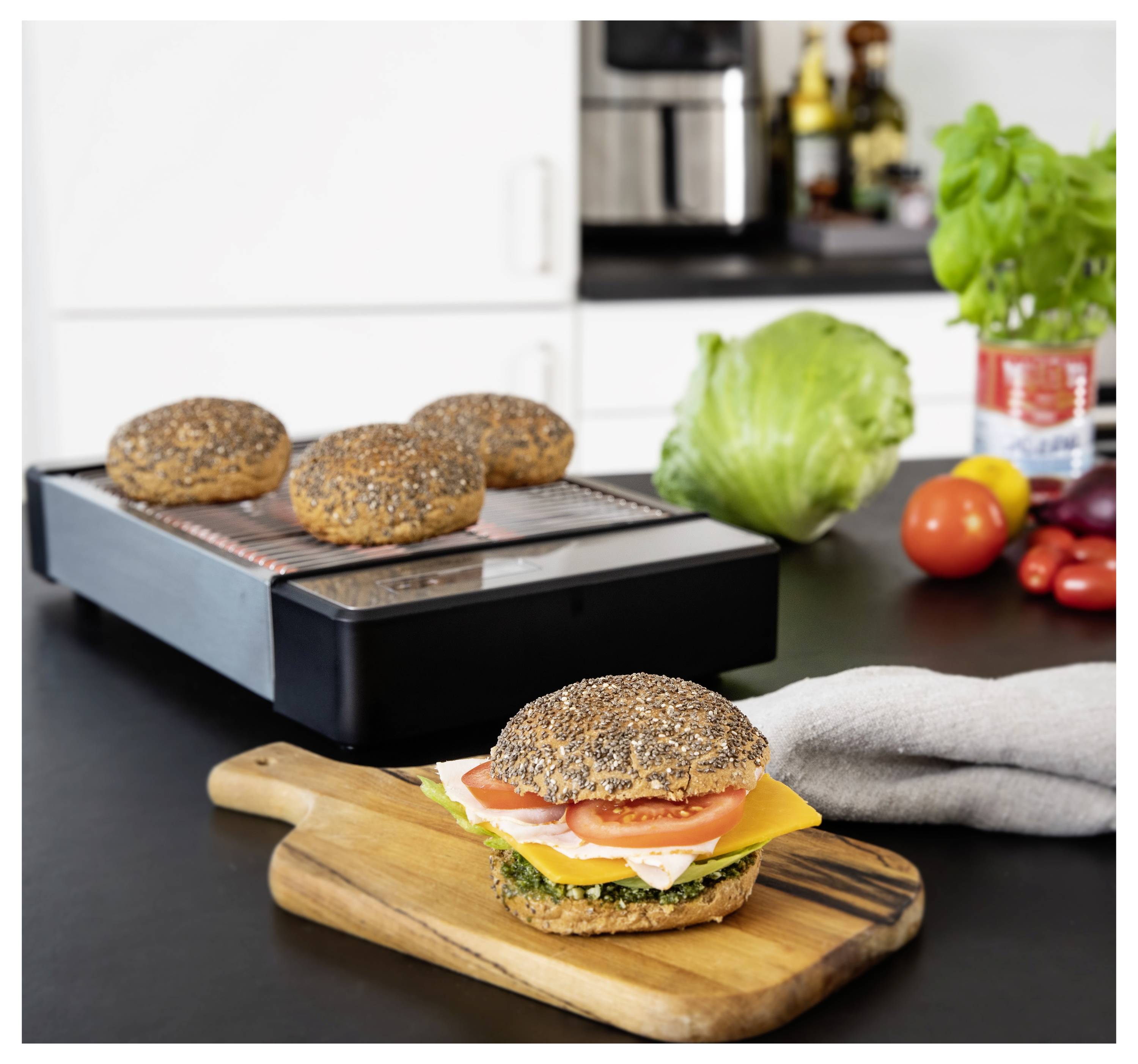 Un sandwich au fromage, aux tomates et à la laitue sur une planche en bois dans une cuisine. Des pains complets sont sur une grille électrique en arrière-plan.