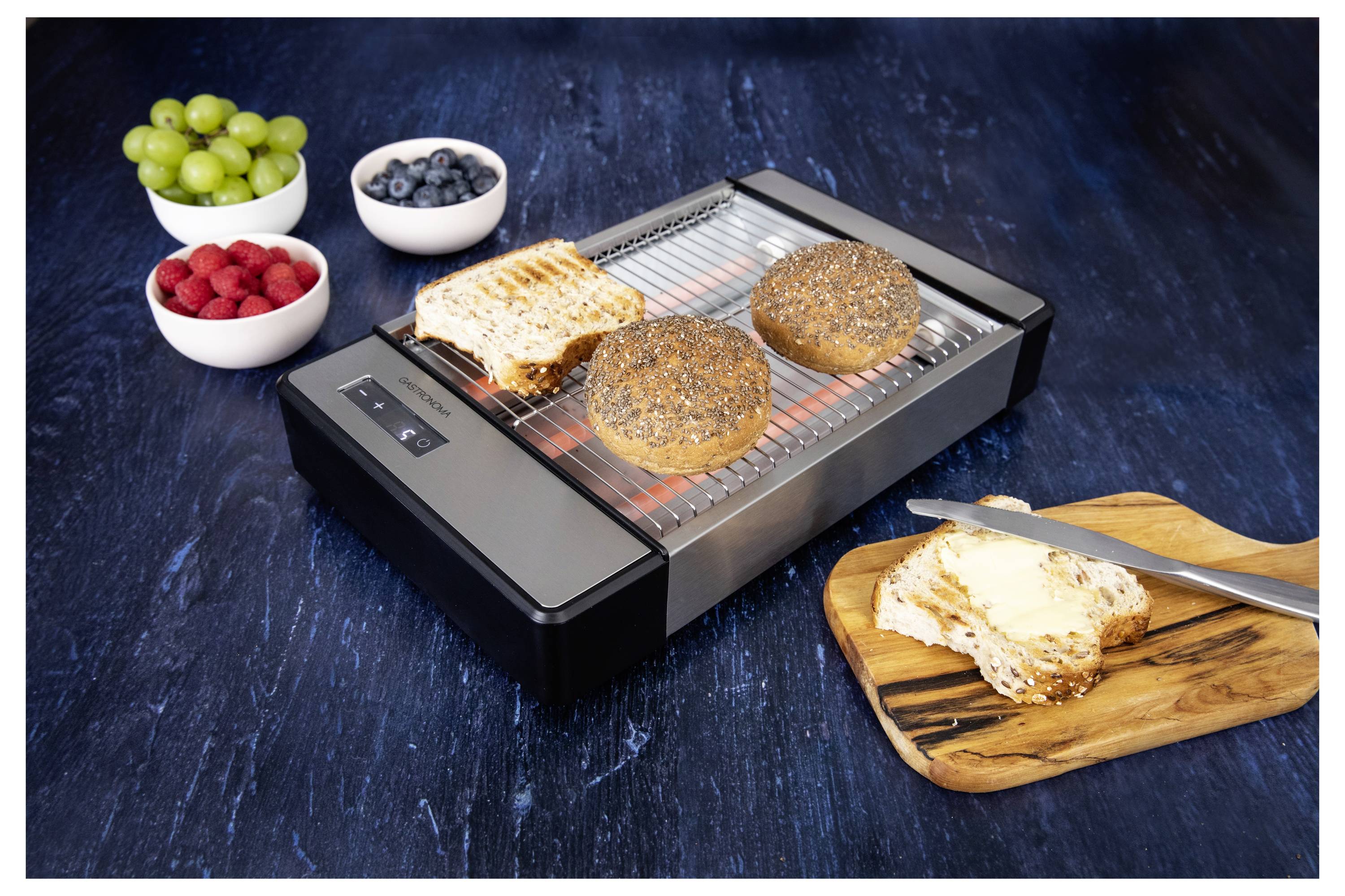 Grille-pain faisant griller du pain et un bagel, couteau sur une tranche beurrée, avec des bols de raisins, de myrtilles et de framboises sur une surface sombre.