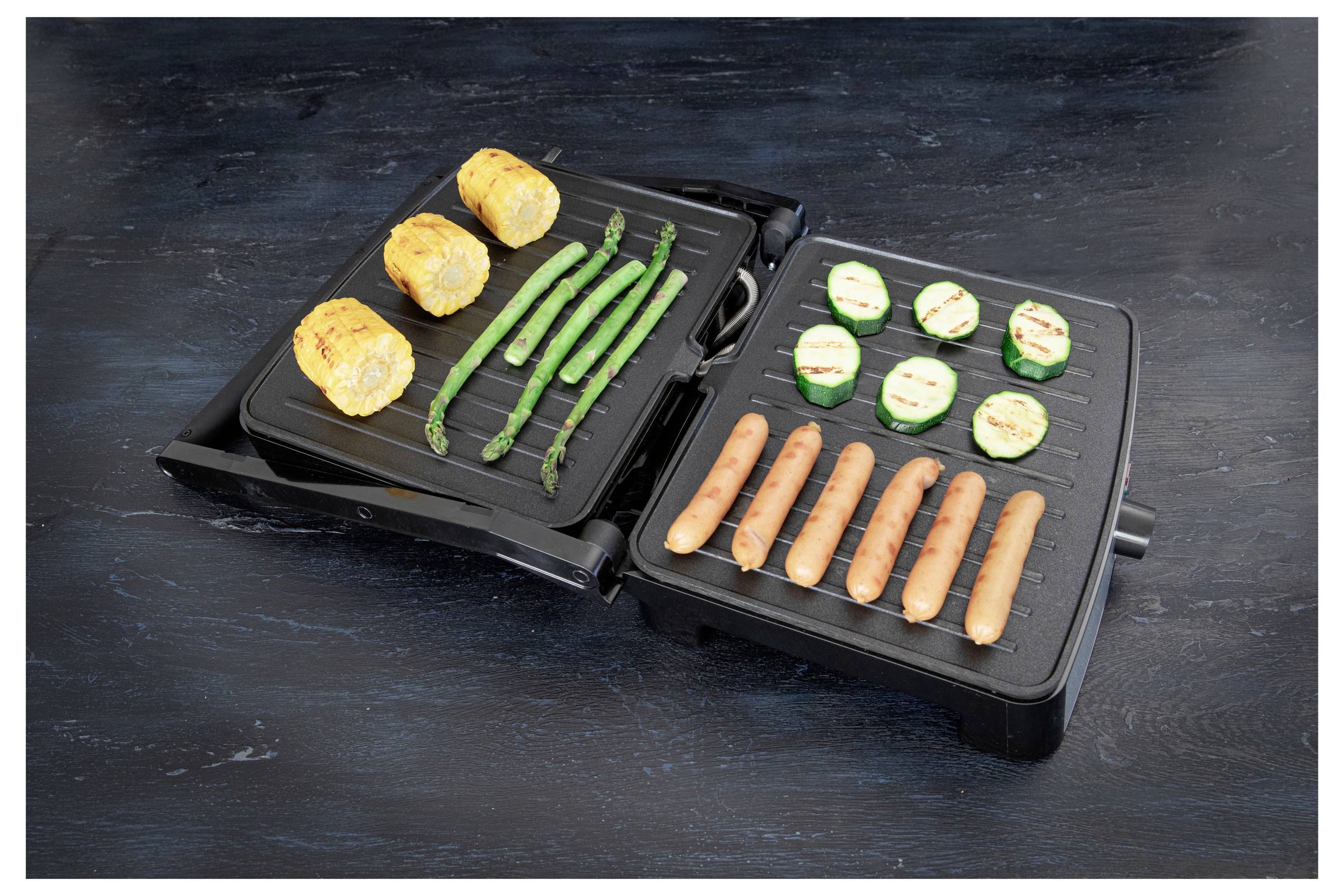 Un grill ouvert avec du maïs grillé, des asperges et des courgettes en tranches d'un côté, des saucisses de l'autre, posé sur une surface sombre.