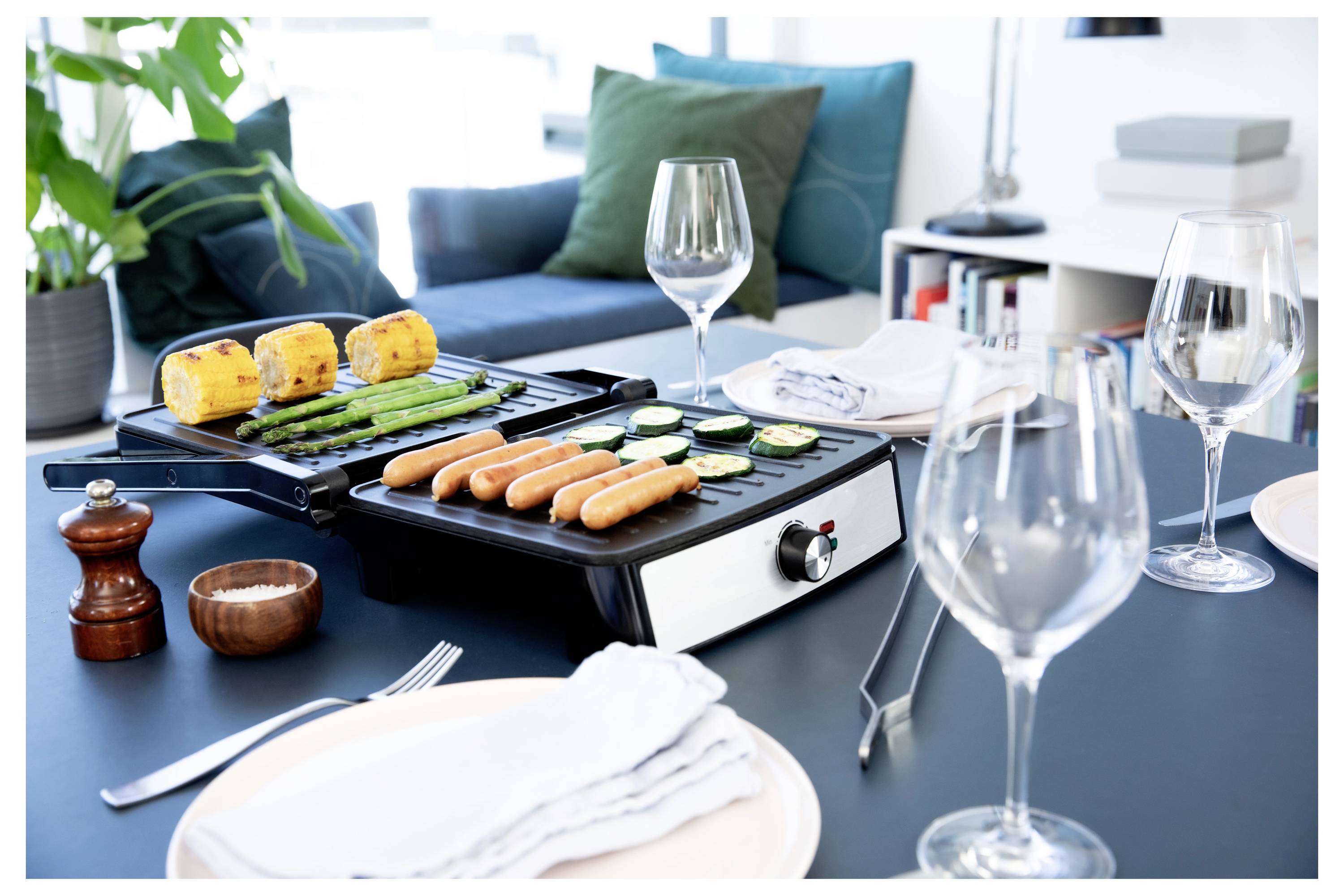 « Une table avec un grill de comptoir cuisinant des saucisses, des courgettes, des asperges et du maïs, disposée à côté de verres à vin, d'assiettes et de serviettes. »