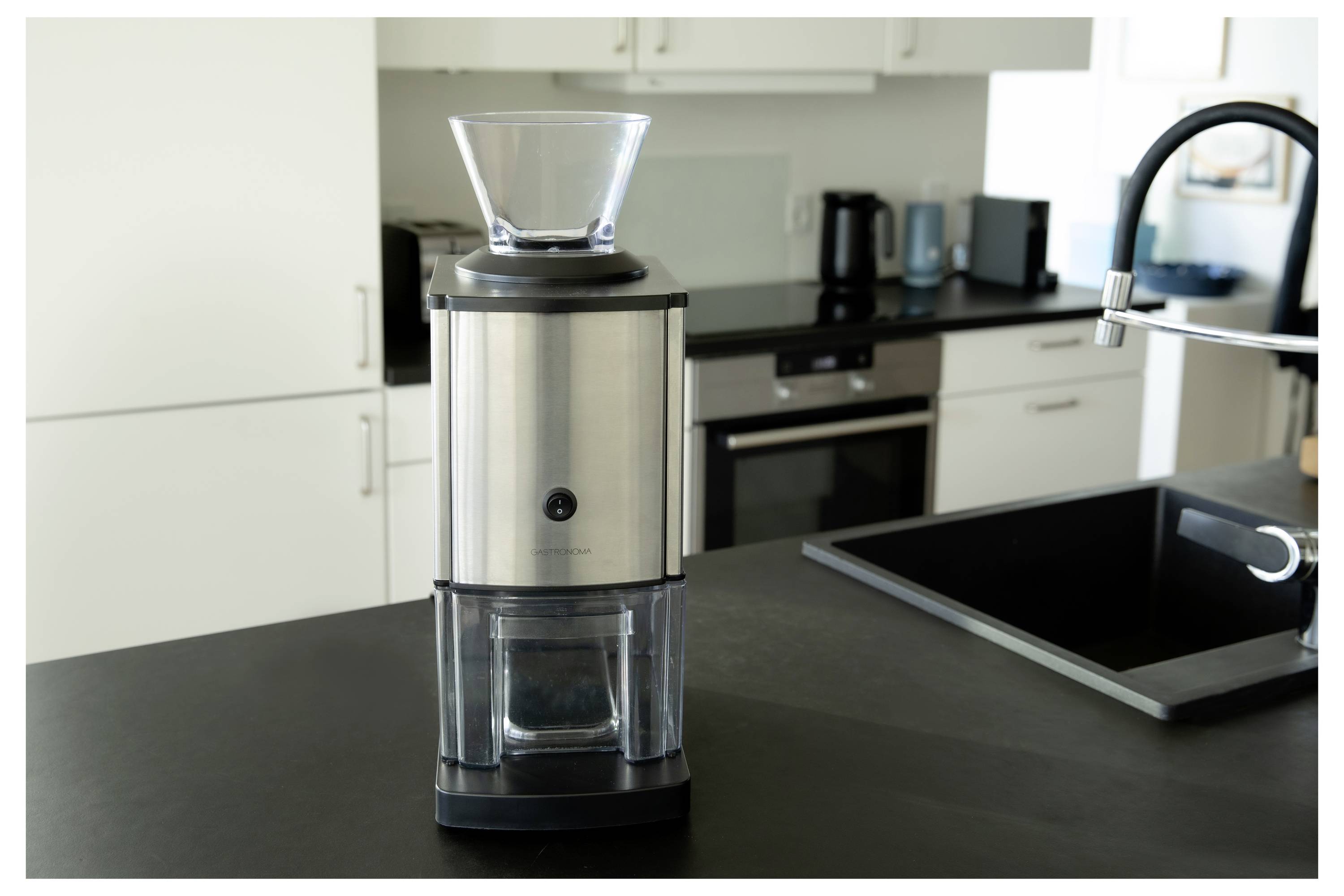 Un moulin à café en acier inoxydable est posé sur un plan de travail de cuisine moderne près d'un évier, avec des armoires blanches et des appareils électroménagers en arrière-plan.