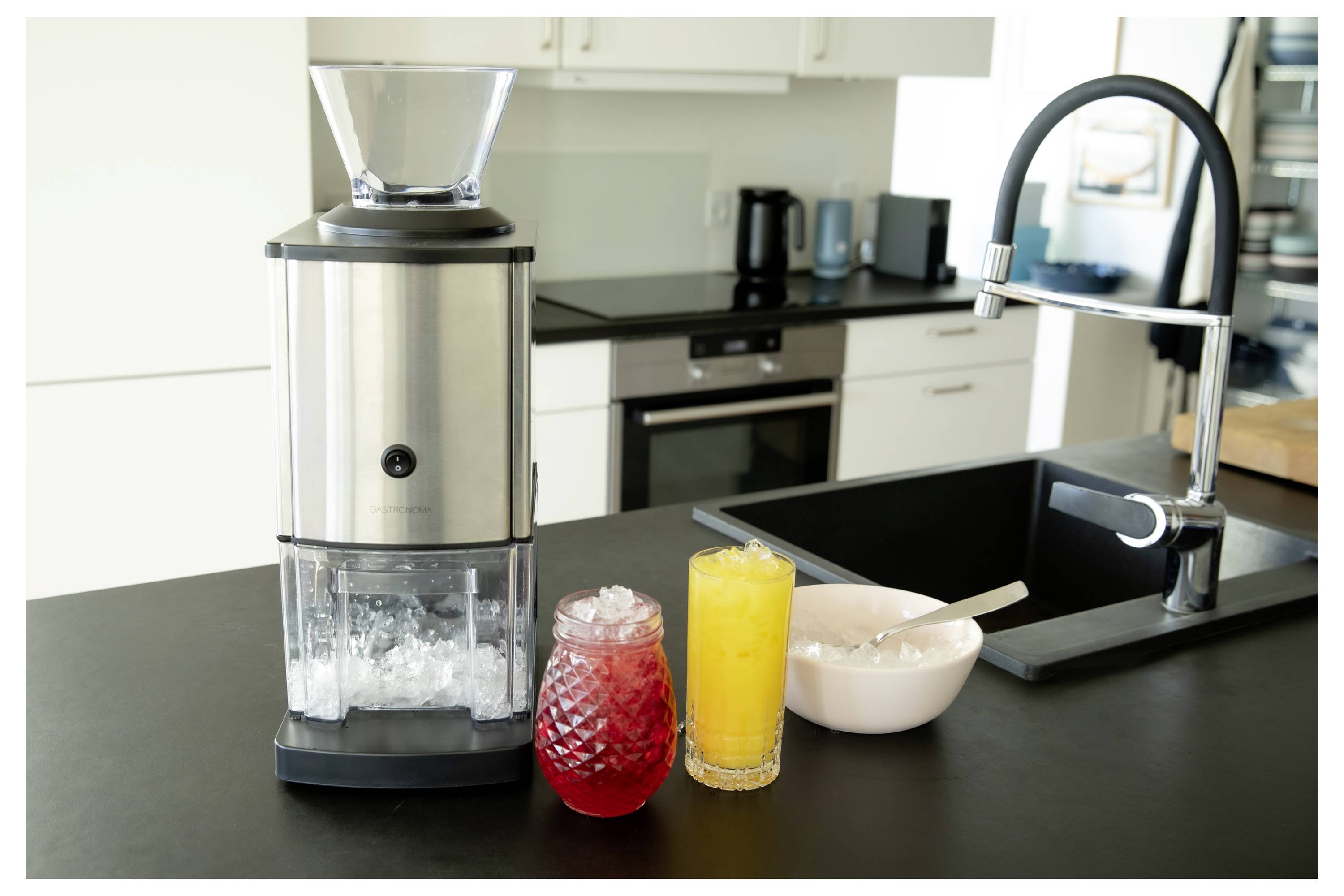 Un comptoir de cuisine avec un broyeur à glace en acier inoxydable, un verre de boisson rouge glacée, un verre de jus d'orange et un bol avec une cuillère à côté d'un évier.