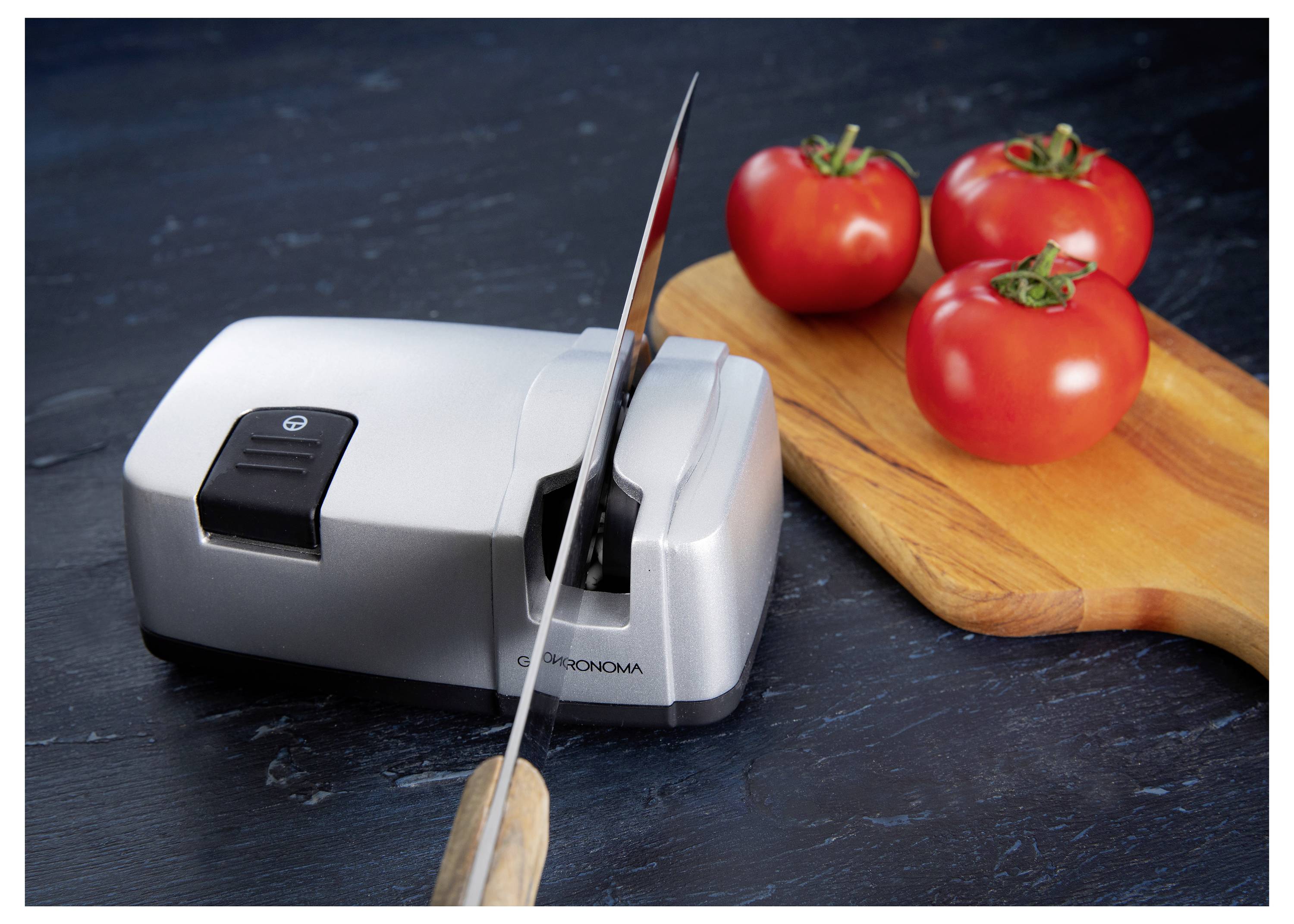 Un couteau est en train d'être aiguisé sur un aiguiseur électrique à côté de trois tomates sur une planche à découper en bois.