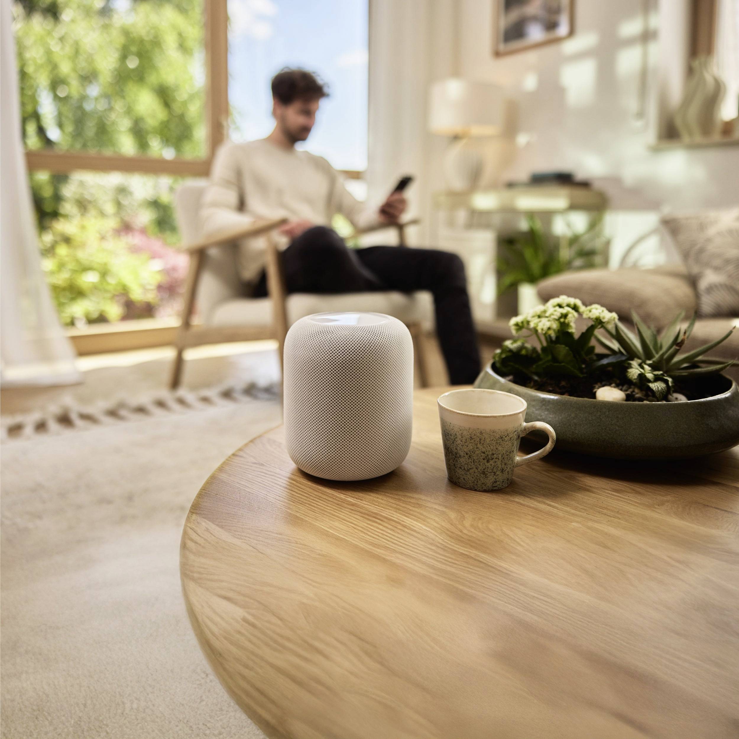 Une personne assise et utilisant un smartphone, avec un haut-parleur intelligent, une tasse à café et une plante sur une table en bois au premier plan.