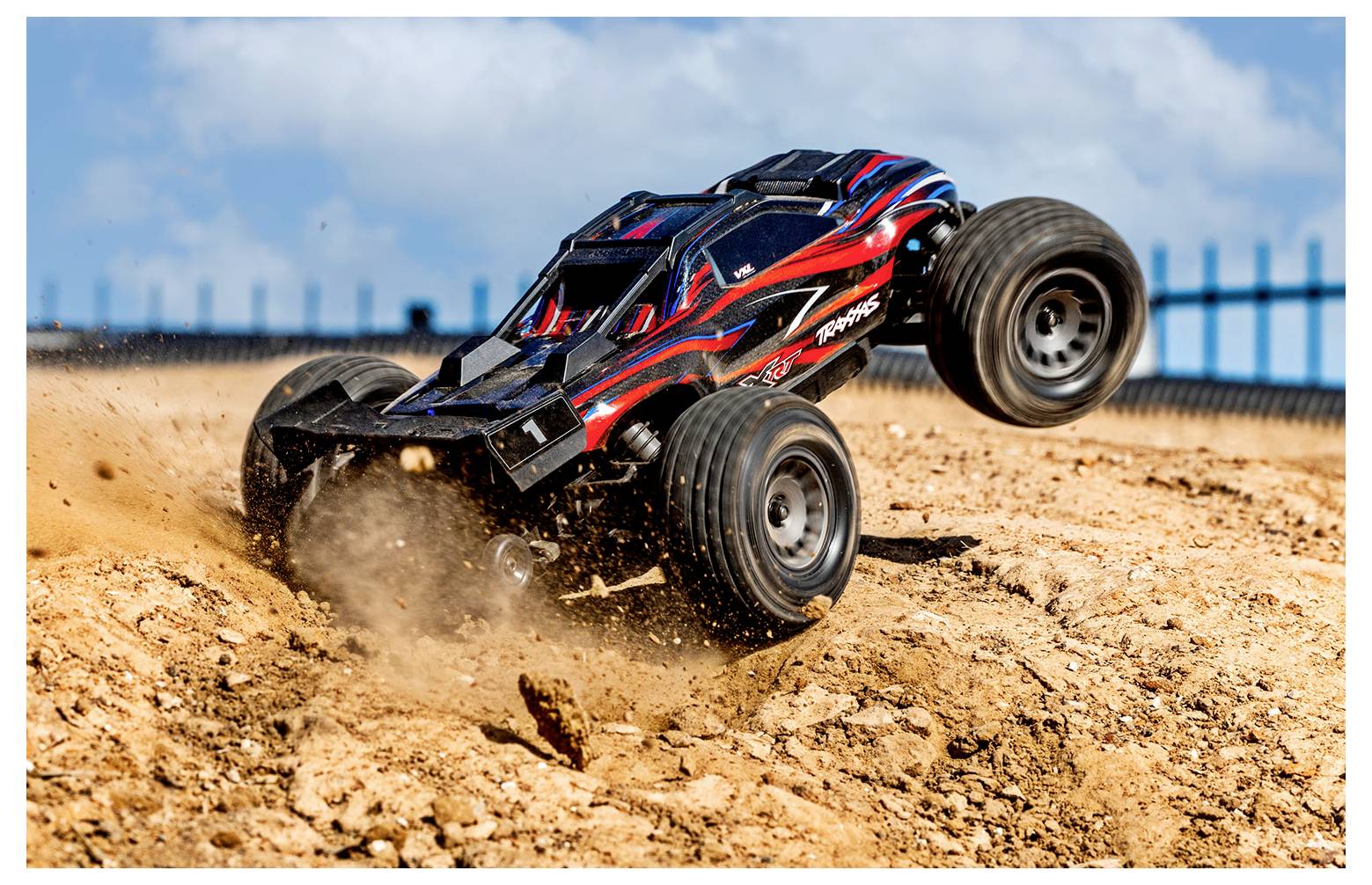 Une voiture télécommandée saute par-dessus un monticule de terre dans un environnement extérieur, soulevant de la poussière sous un ciel bleu parsemé de quelques nuages.