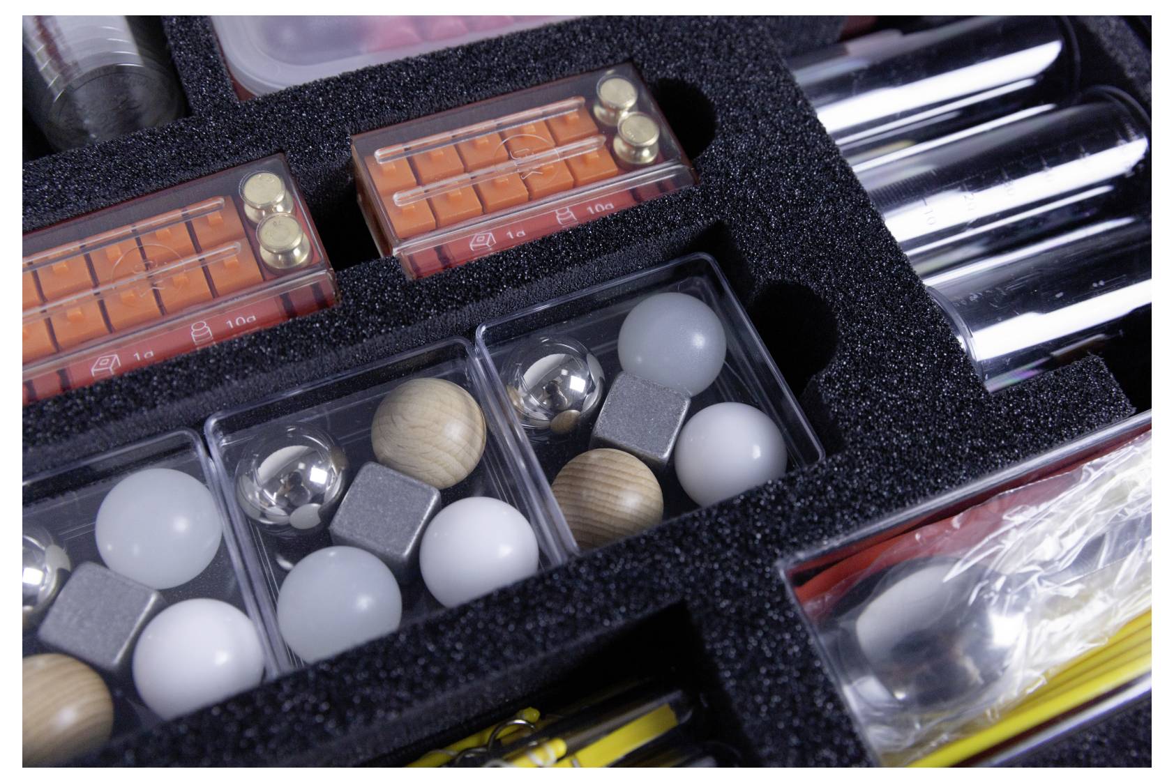 A foam-lined case containing metallic and wooden spheres, rectangular magnets, and transparent containers, likely part of a science kit.