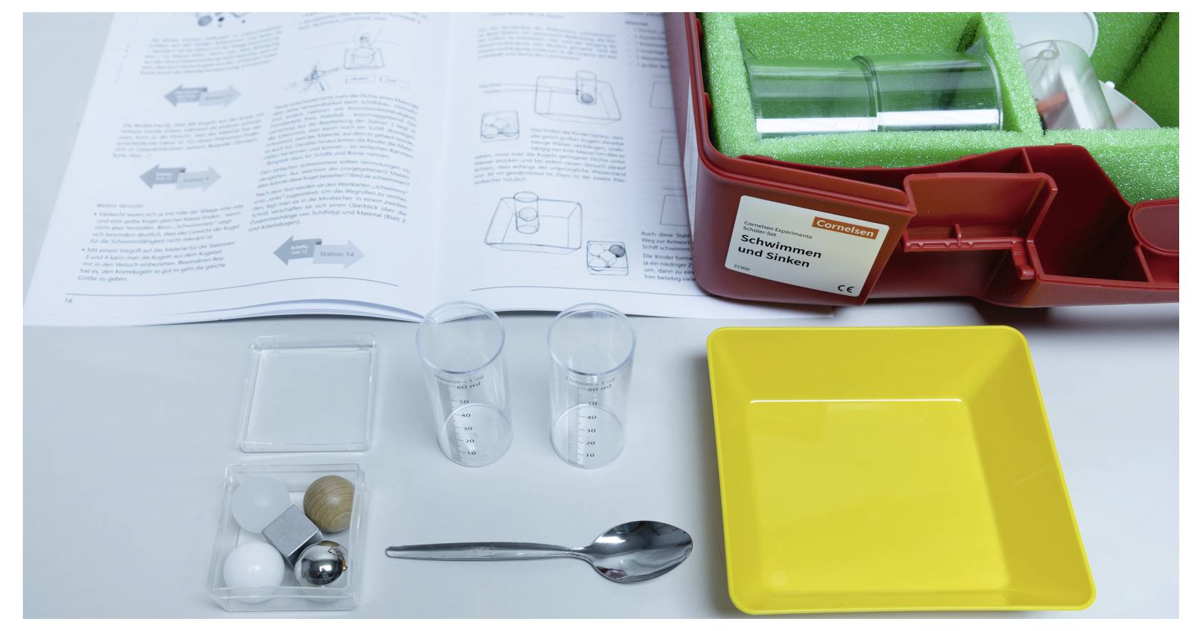 A science kit includes beakers, a spoon, a yellow tray, various items like balls and cubes, and an open instruction manual on a table.
