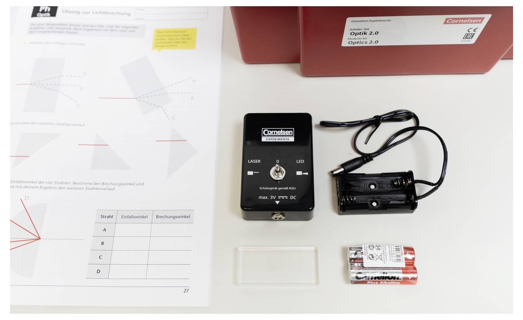 'A desk with a laser module, battery holder with AA batteries, a small rectangular object, and an optics worksheet labeled 