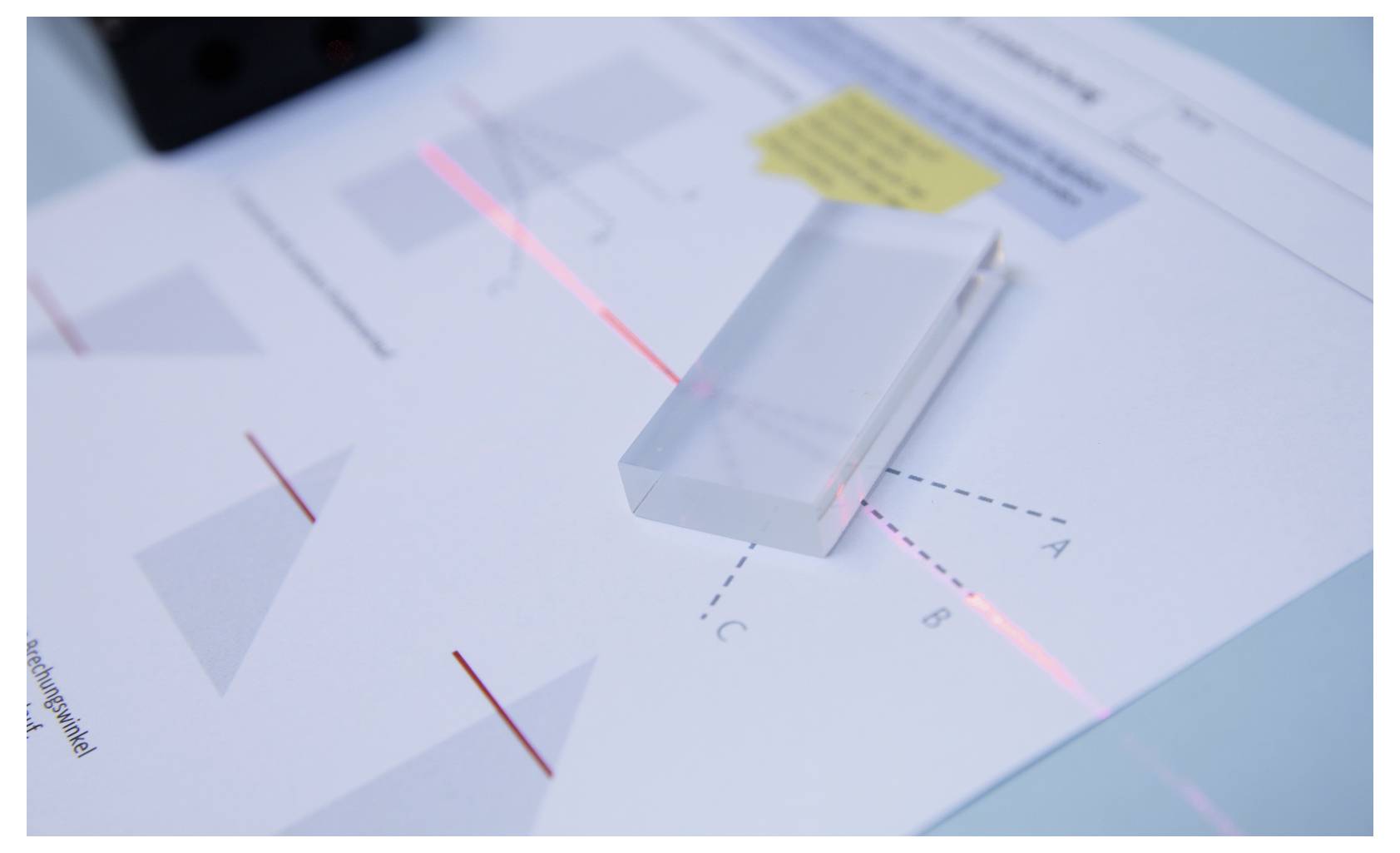A beam of light passes through a transparent prism, refracting into different paths marked A, B, and C on a worksheet underneath.