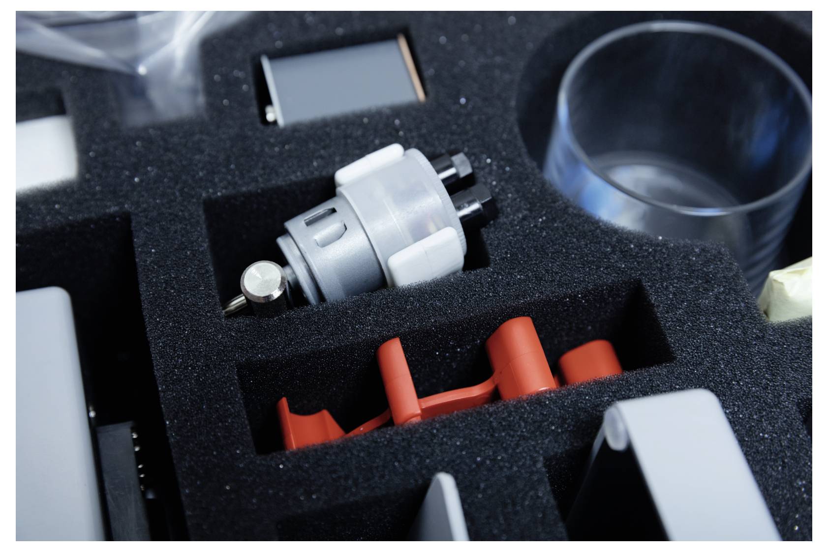 Close-up of a plastic case with various organized tools, including a silver cylindrical part and a red handle, in foam compartments.