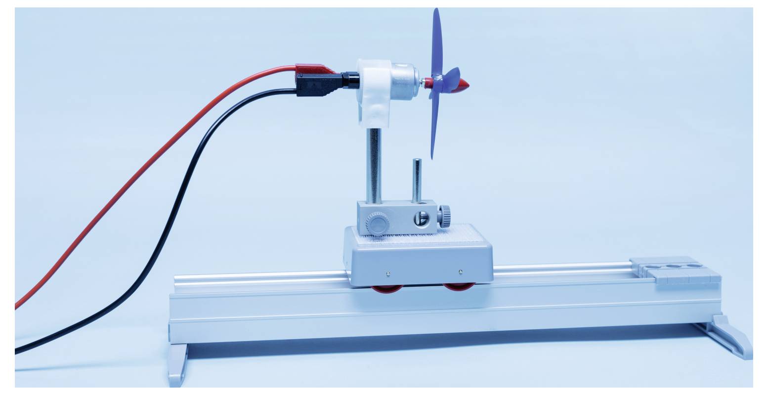 A small wind turbine model with red and black wires, mounted on a metallic base, set against a light blue background.