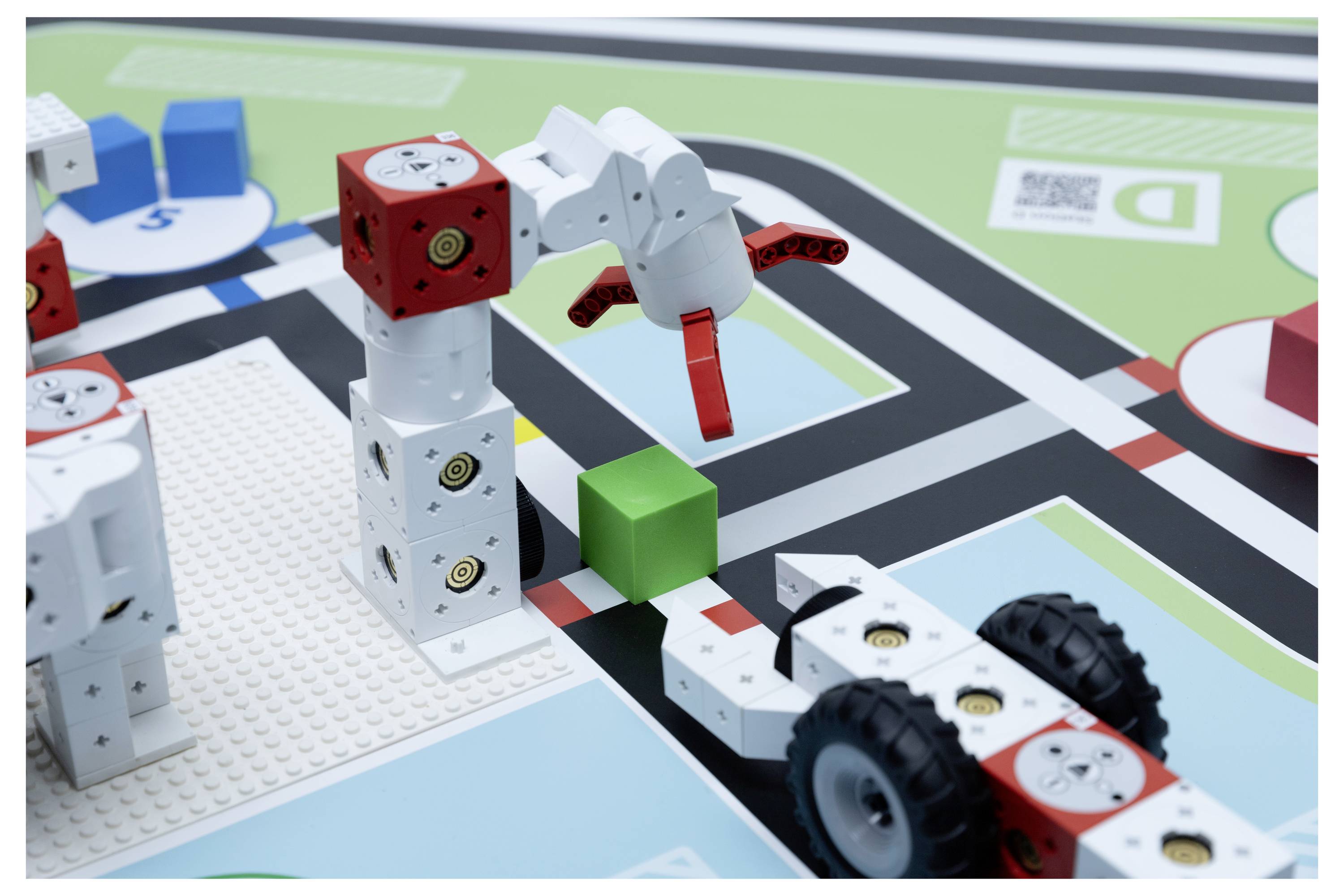 A robotic arm stacking white and red blocks on a board with roads, near a wheeled vehicle and a green cube.