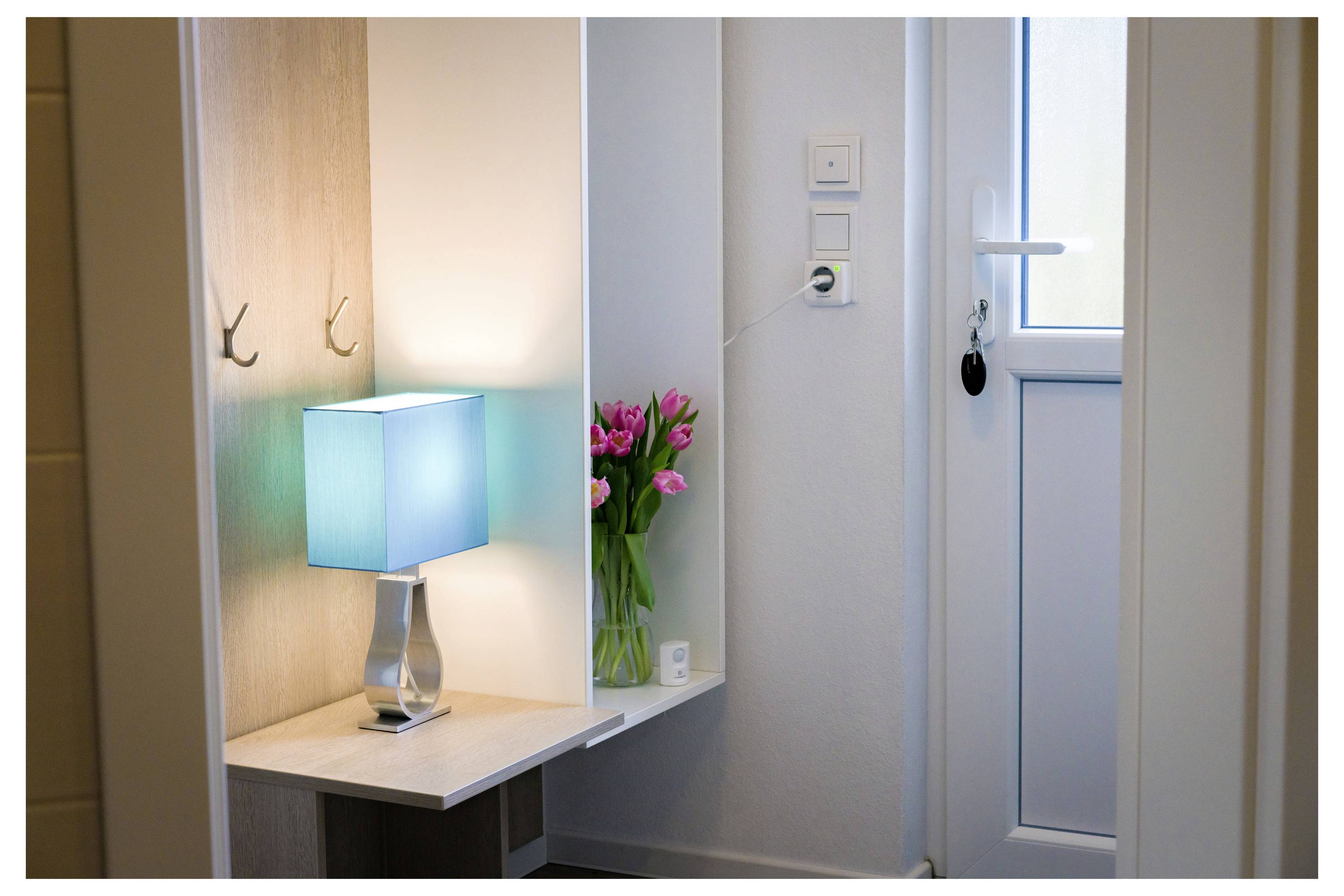 Un petit couloir avec une lampe turquoise sur une table d'angle, des tulipes roses dans un vase, et une porte blanche fermée avec une clé dans la serrure.