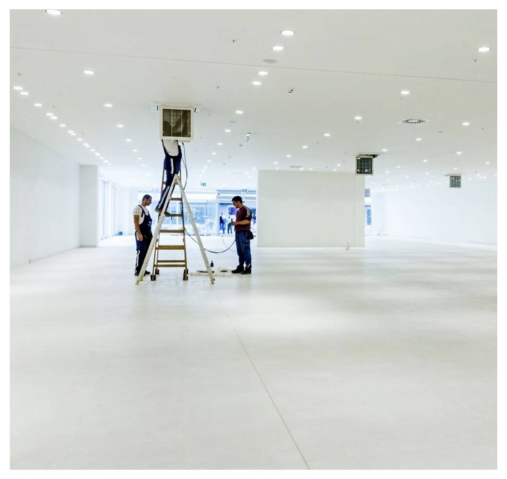 Deux travailleurs installant ou vérifiant une unité de plafond dans une grande pièce vide, bien éclairée, avec un plafond haut et un design minimaliste.