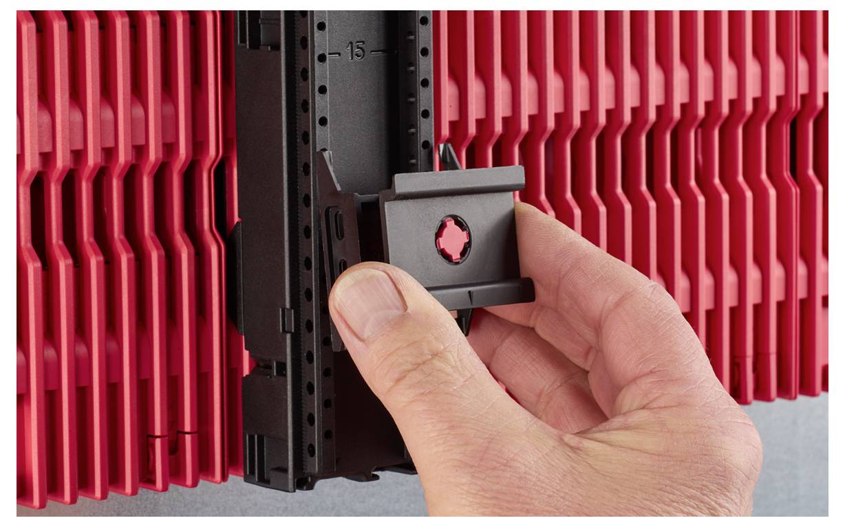 Une main insérant un clip en plastique noir sur une surface verticale rouge rainurée, probablement un système de rangement ou d'organisation.