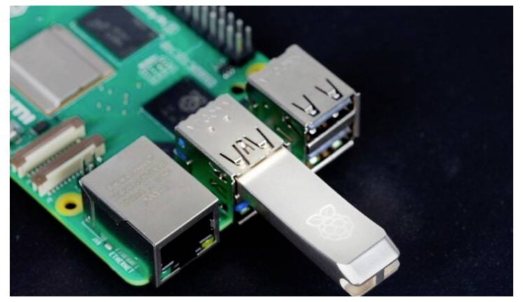A USB drive inserted into a Raspberry Pi board showing USB ports and Ethernet connection, resting on a dark surface.