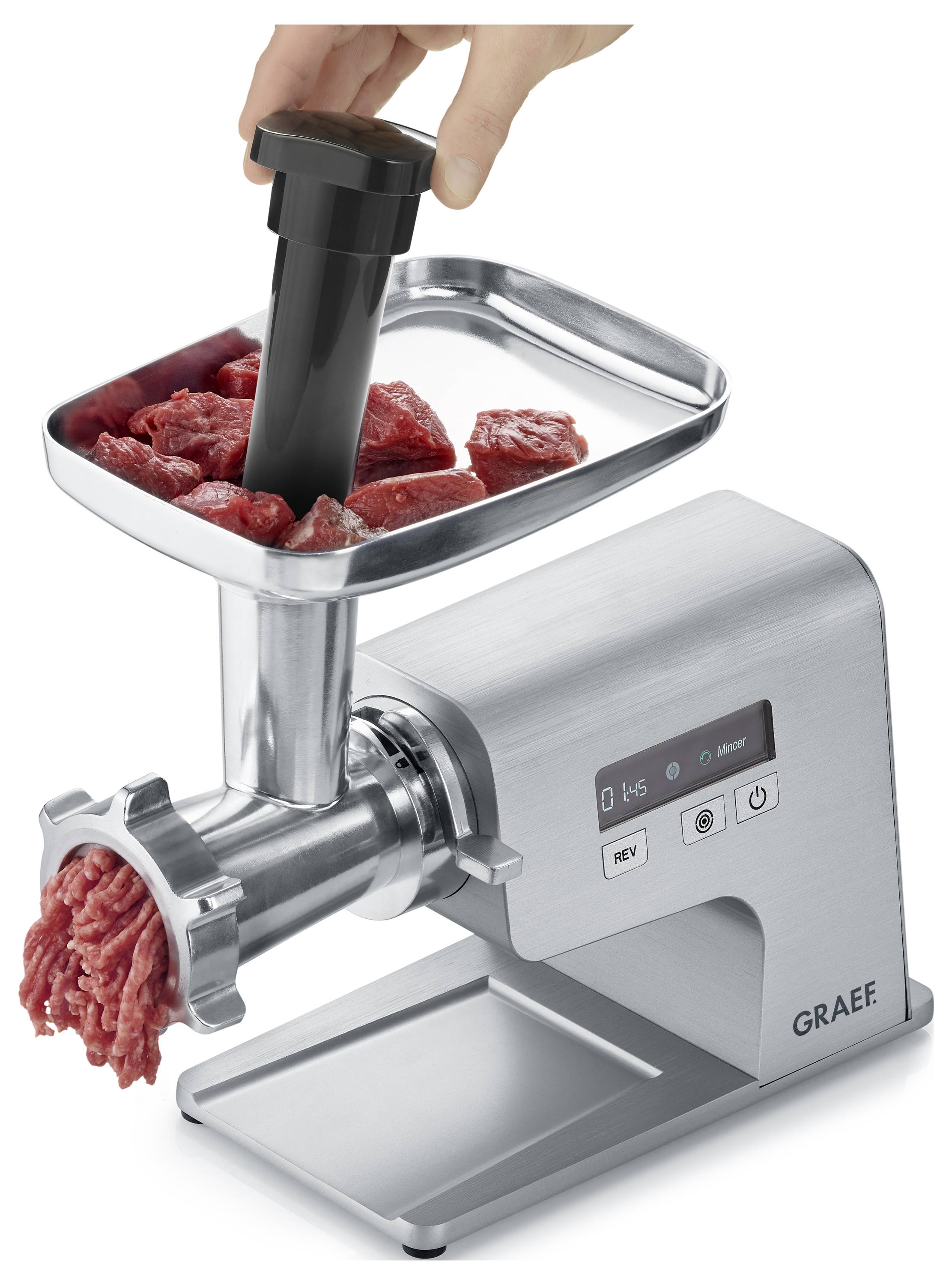 Un hachoir électrique argenté avec un plateau sur le dessus contenant de la viande crue. Une main utilise un poussoir pour introduire la viande dans le hachoir, produisant de la viande hachée.