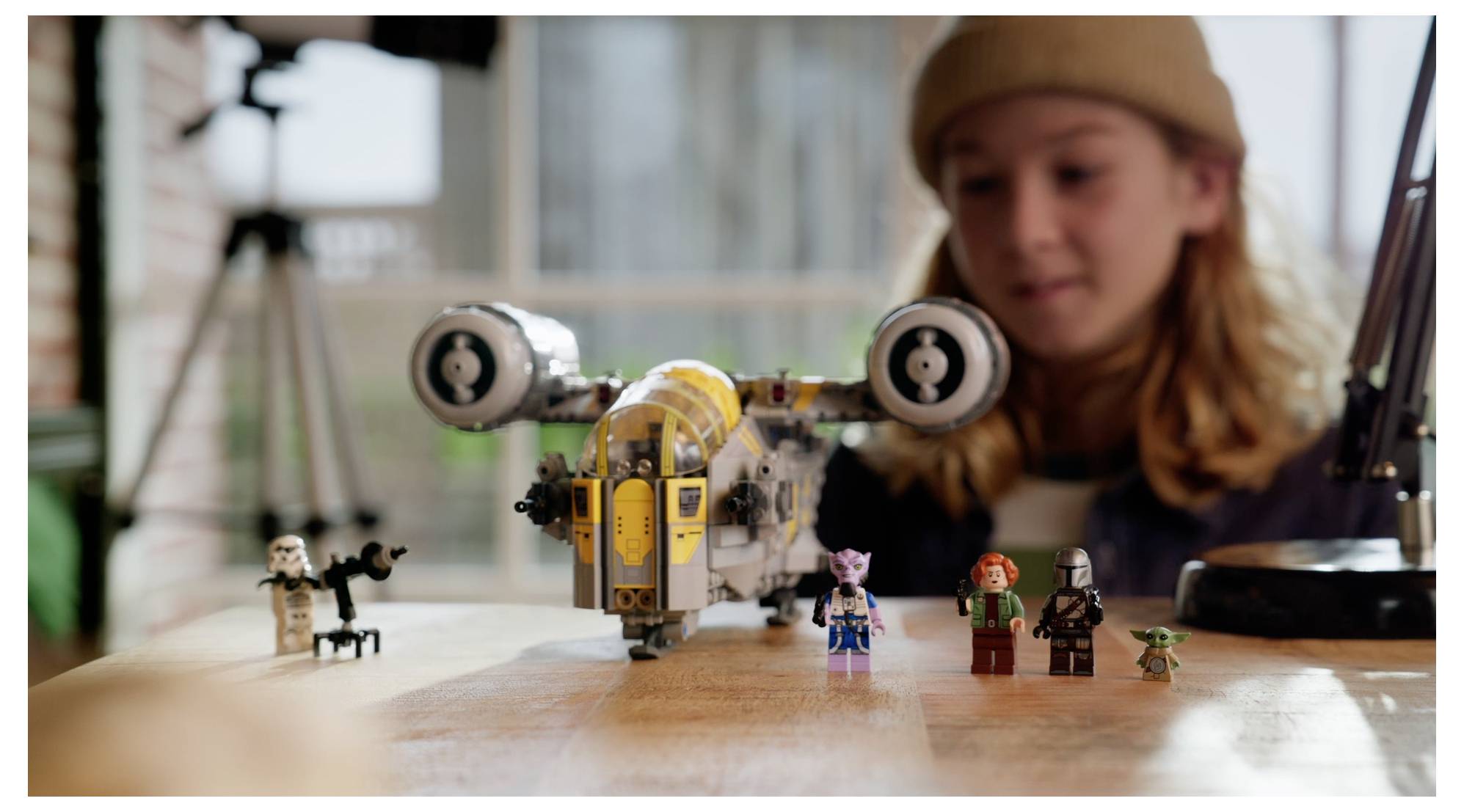 Un enfant se concentre sur l'assemblage d'un modèle détaillé de vaisseau spatial avec de petites figurines de personnages, sur une table en bois, dans une pièce lumineuse.