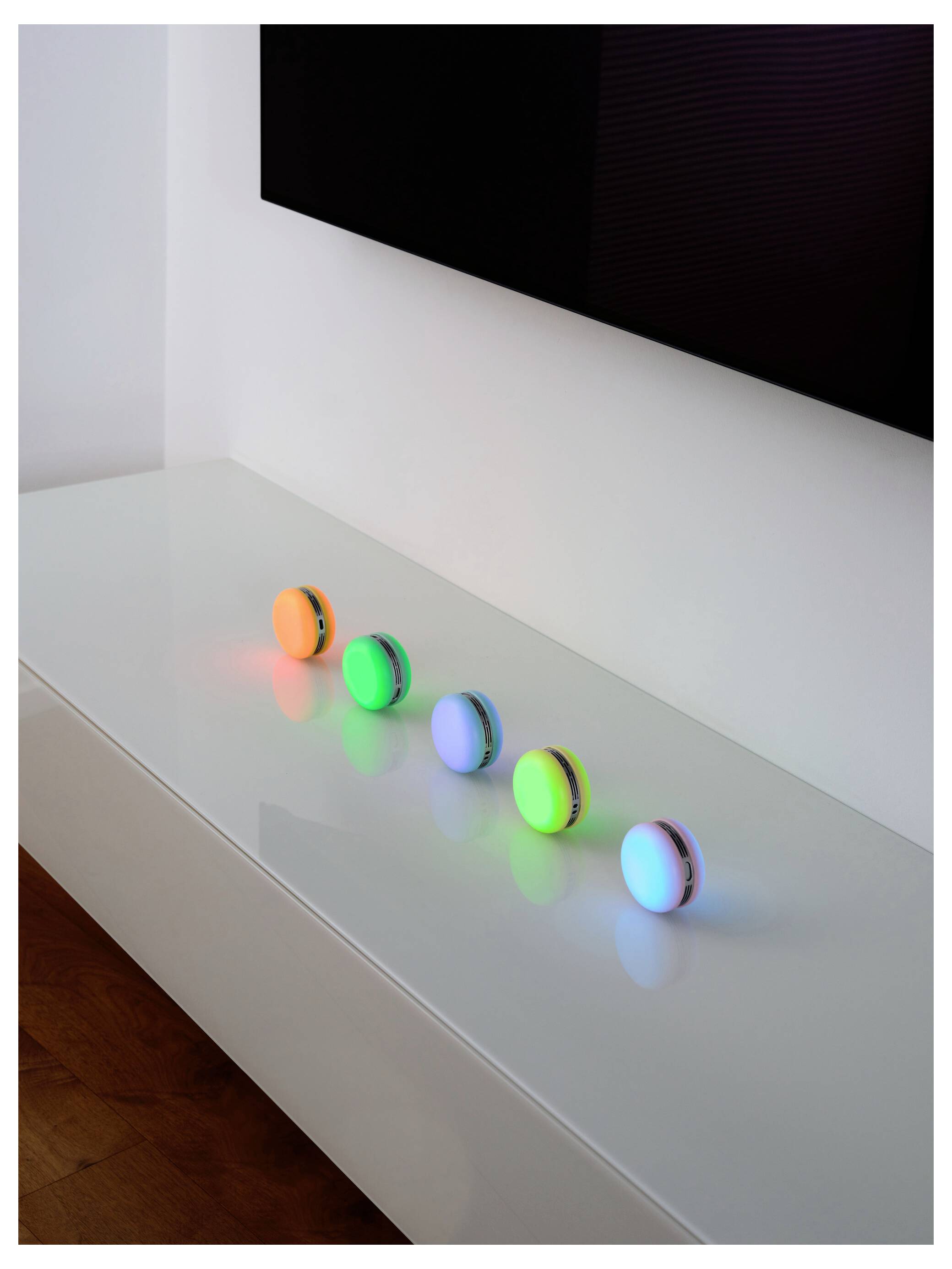 Cinq dispositifs circulaires colorés et lumineux sur une surface blanche réfléchissante. Chaque dispositif possède une couleur de lumière différente : orange, vert, bleu, violet et rouge.